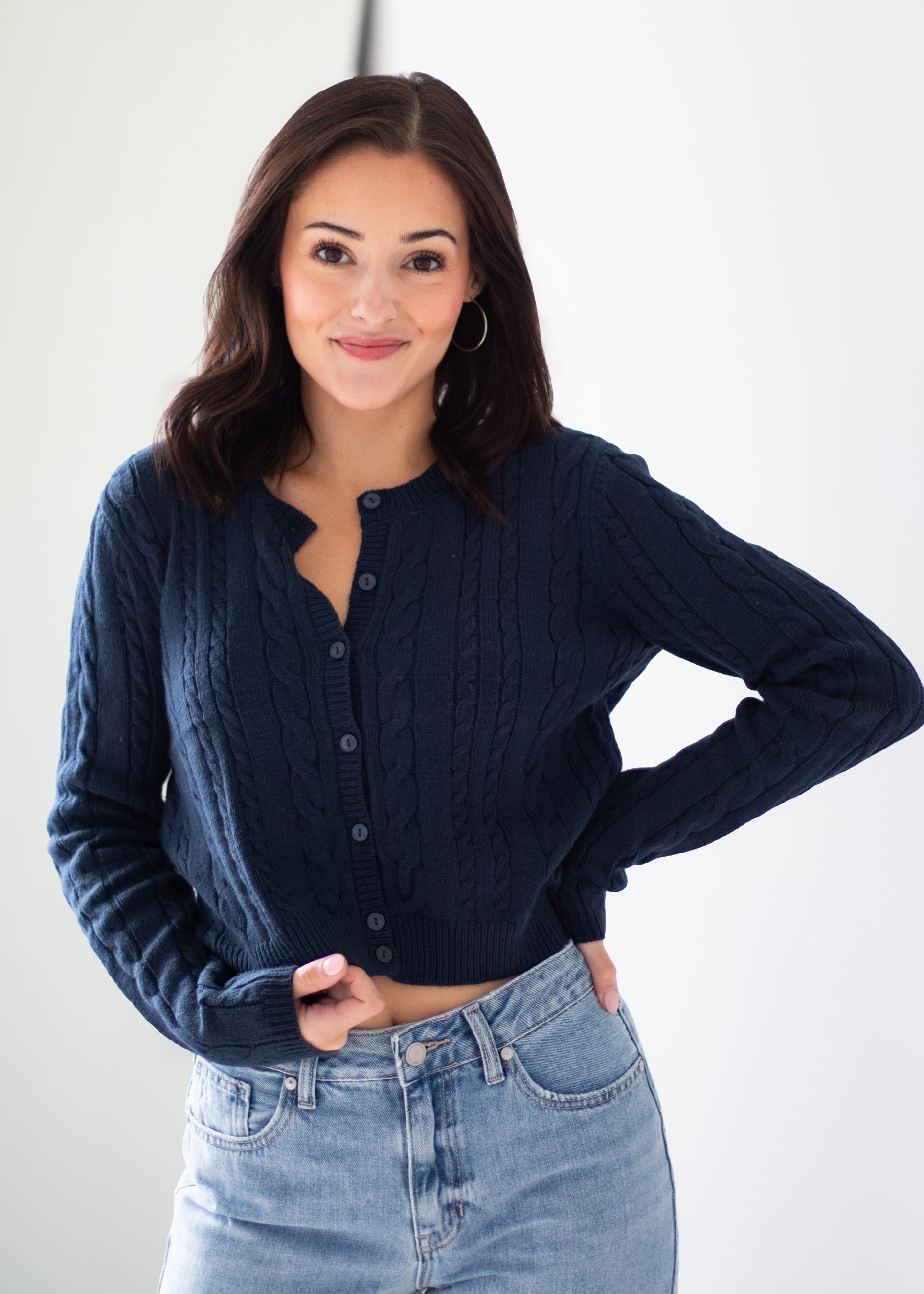 A woman wearing a navy blue cable-knit button-up cardigan sweater and light-wash blue jeans, posing against a clean white studio background.