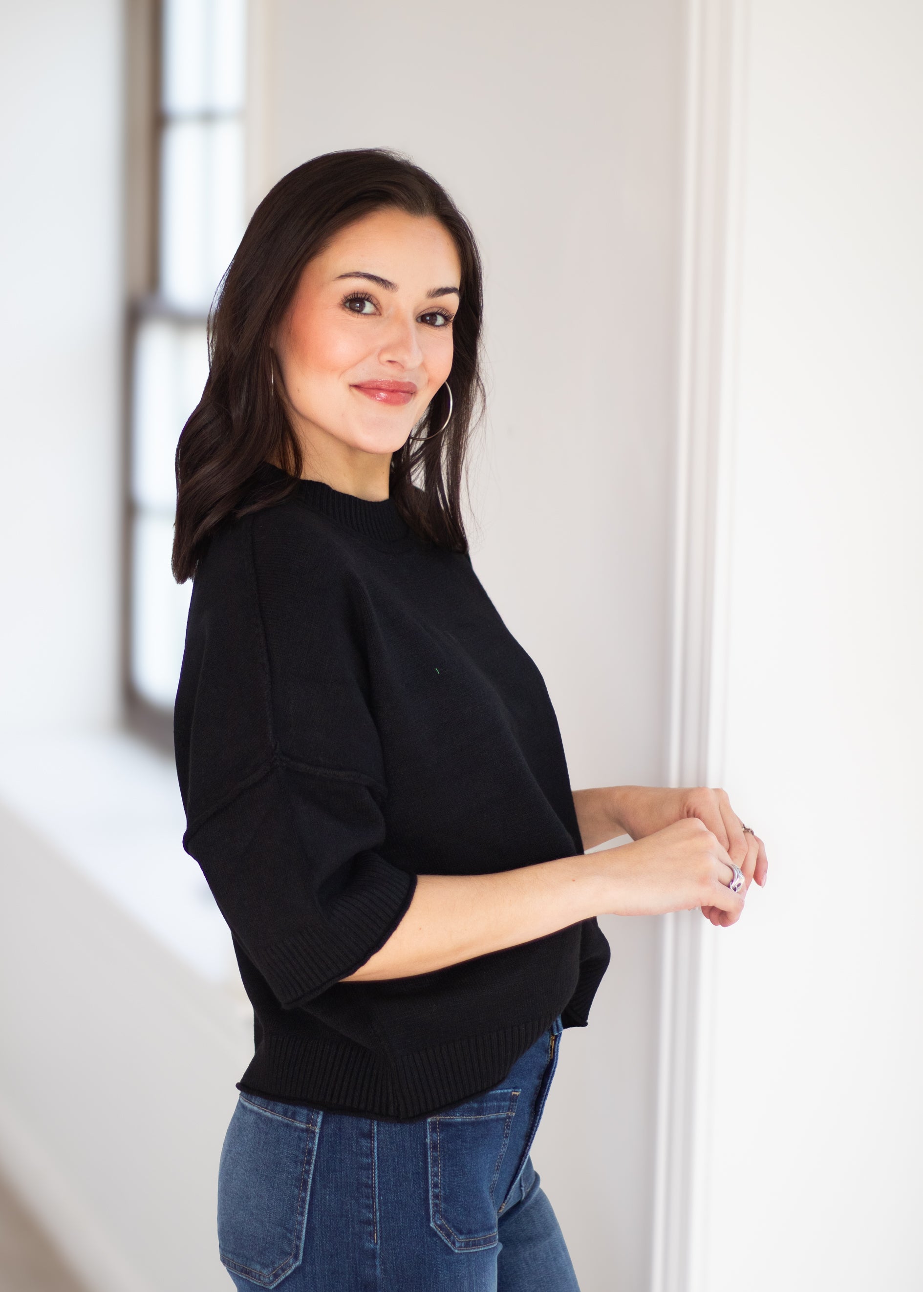 A woman with dark hair wearing a black knit sweater and blue jeans, standing in a brightly lit room with white walls.