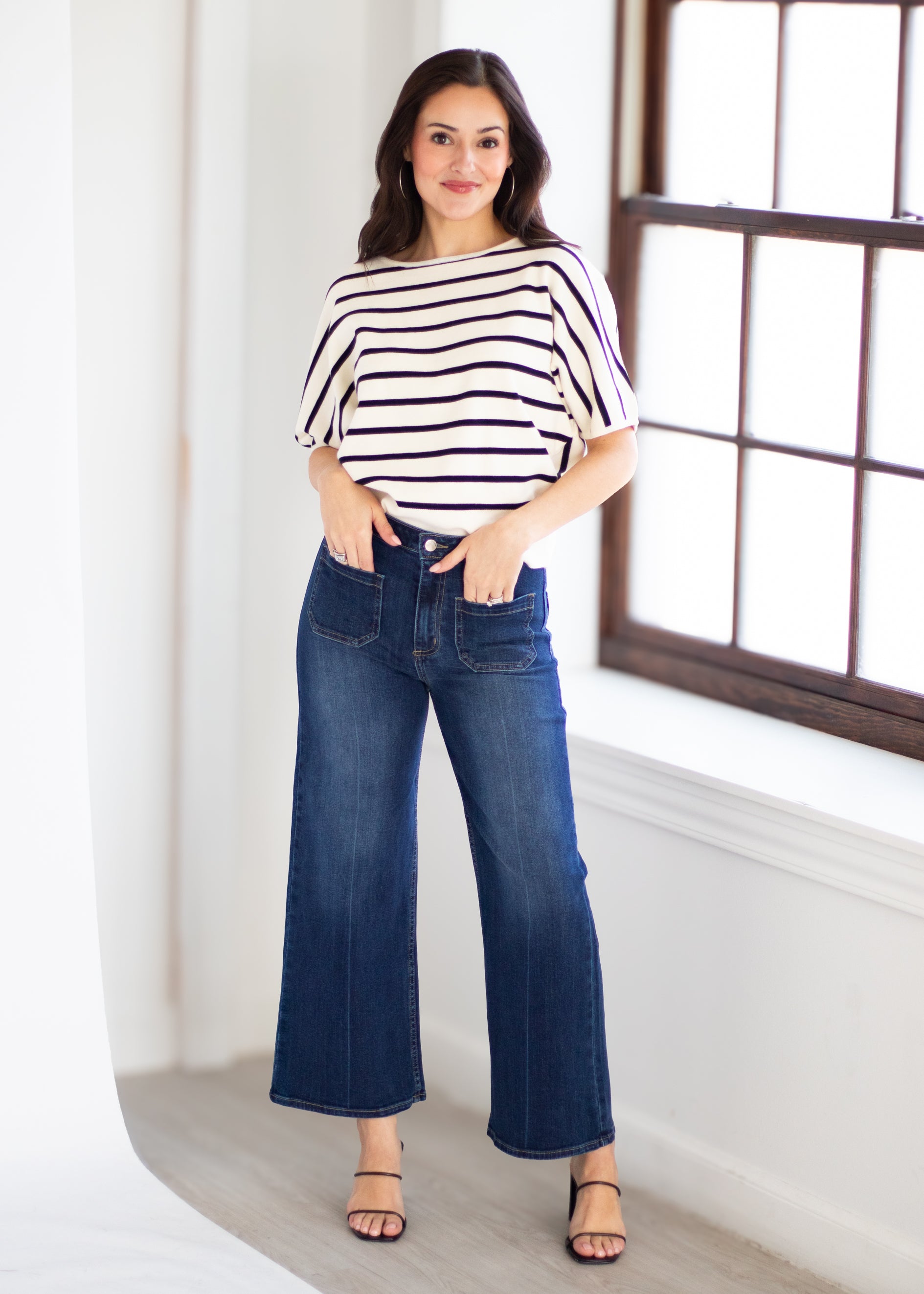 A woman modeling a white short-sleeve top with thin navy horizontal stripes and dark blue wide-leg cropped jeans, standing in a bright room with a window.