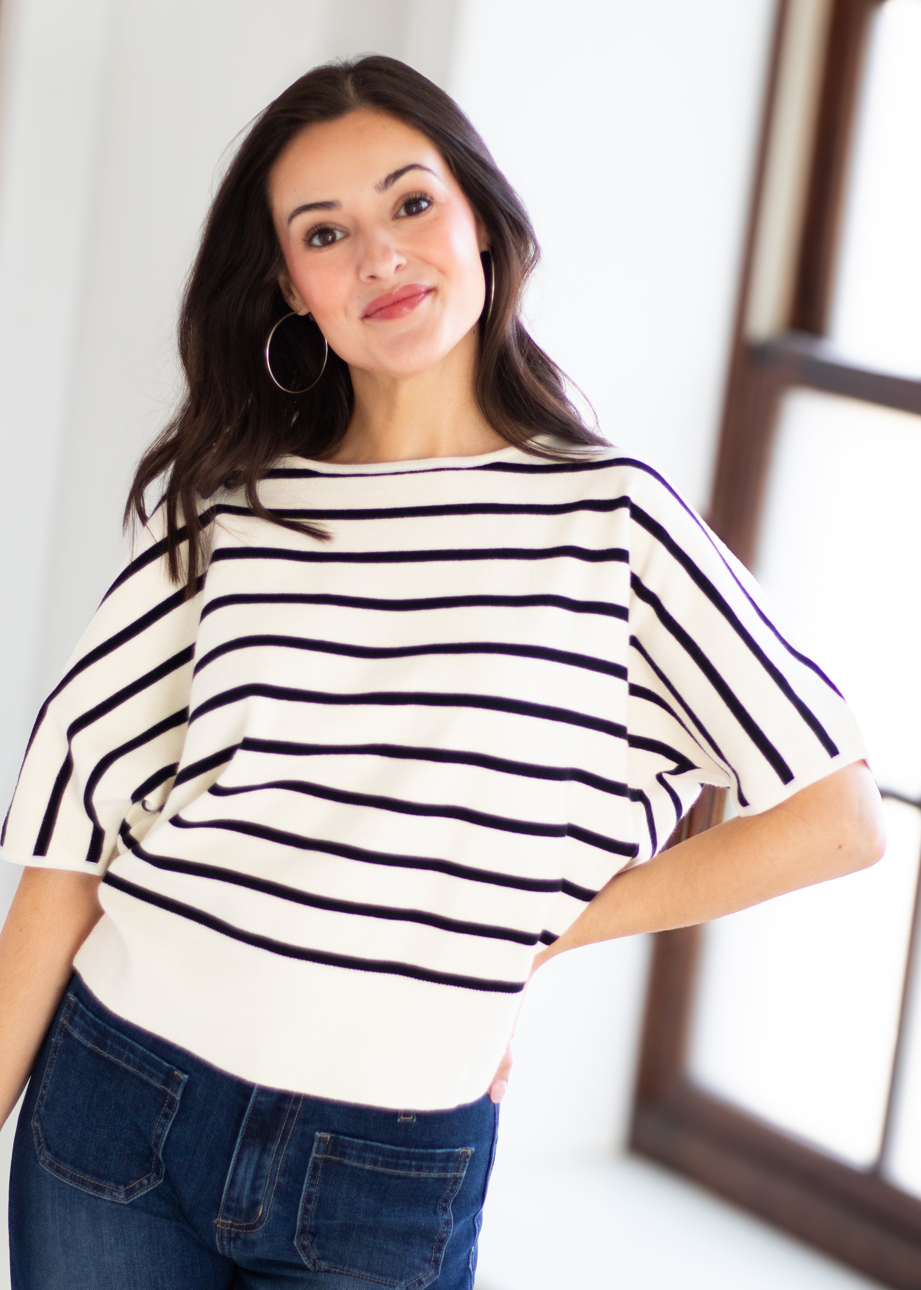 A woman wearing a cream knit top with thin black horizontal stripes and blue jeans, standing in a brightly lit indoor setting.