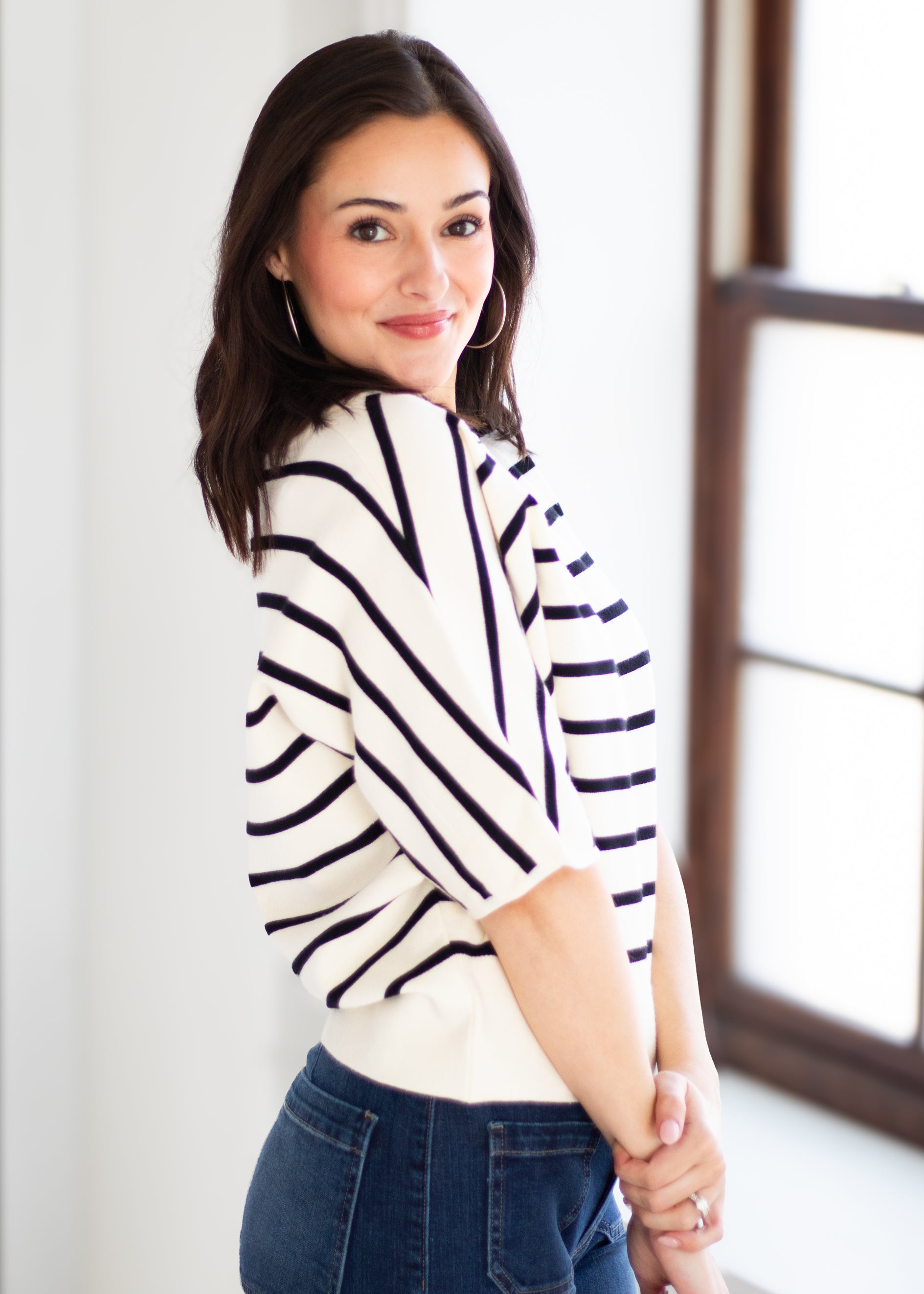 A woman wearing a cream knit sweater with navy blue diagonal stripes and blue jeans, posing in a bright indoor setting with a window in the background.