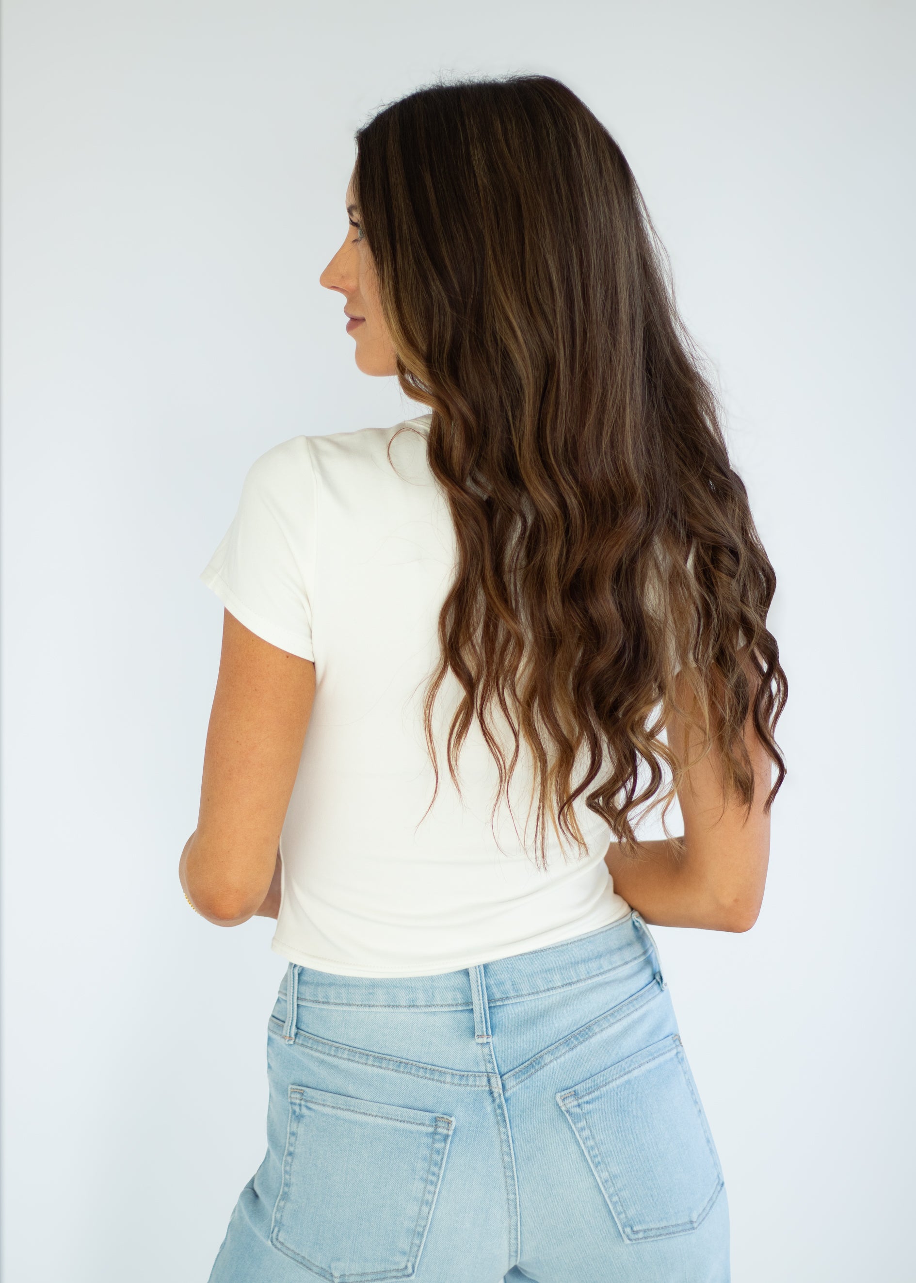 Woman with long wavy hair wearing a white t-shirt and light blue jeans against a plain background