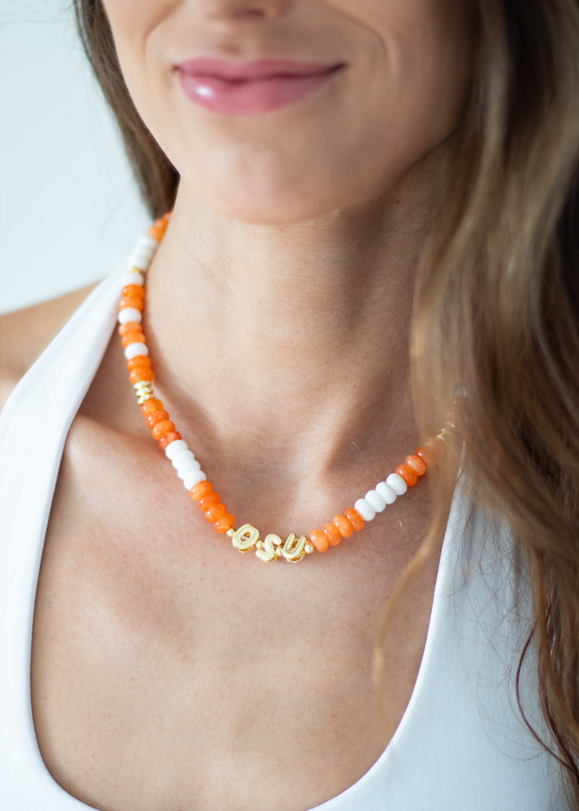 A close-up of a woman wearing a beaded necklace with orange and white beads and gold letter charms spelling 'SOUL'.