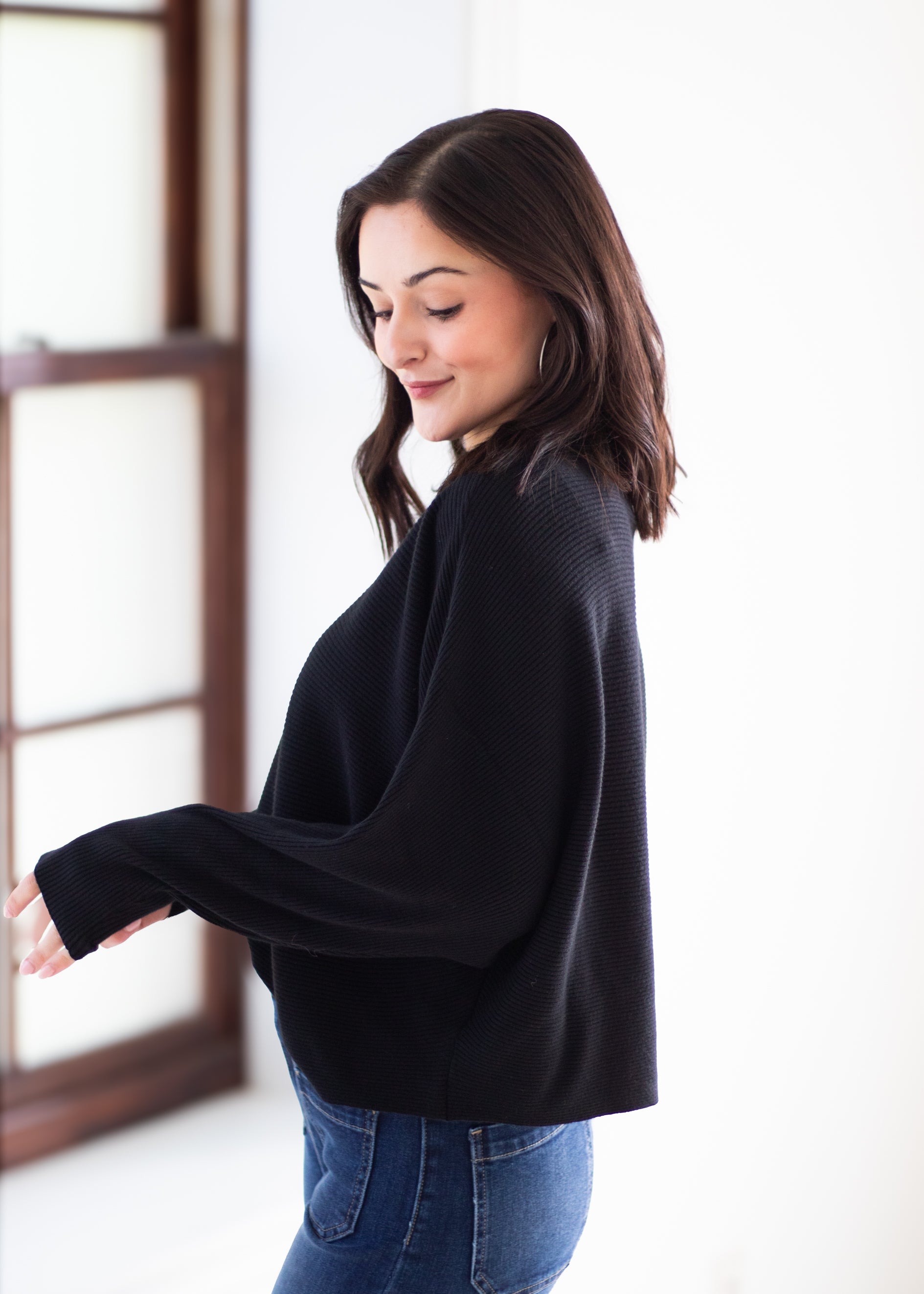 A woman wearing a black textured knit long-sleeve sweater and blue jeans, standing in a bright indoor setting.