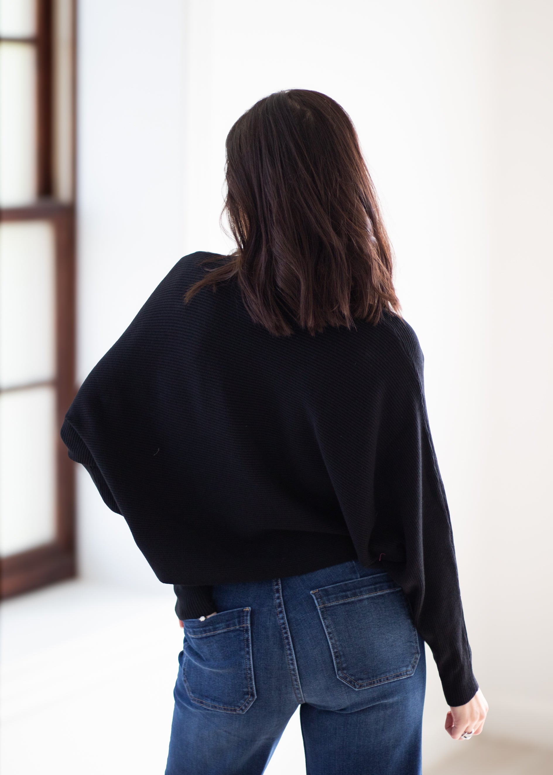 A woman viewed from behind wearing a black knit sweater and blue jeans in a bright indoor setting.