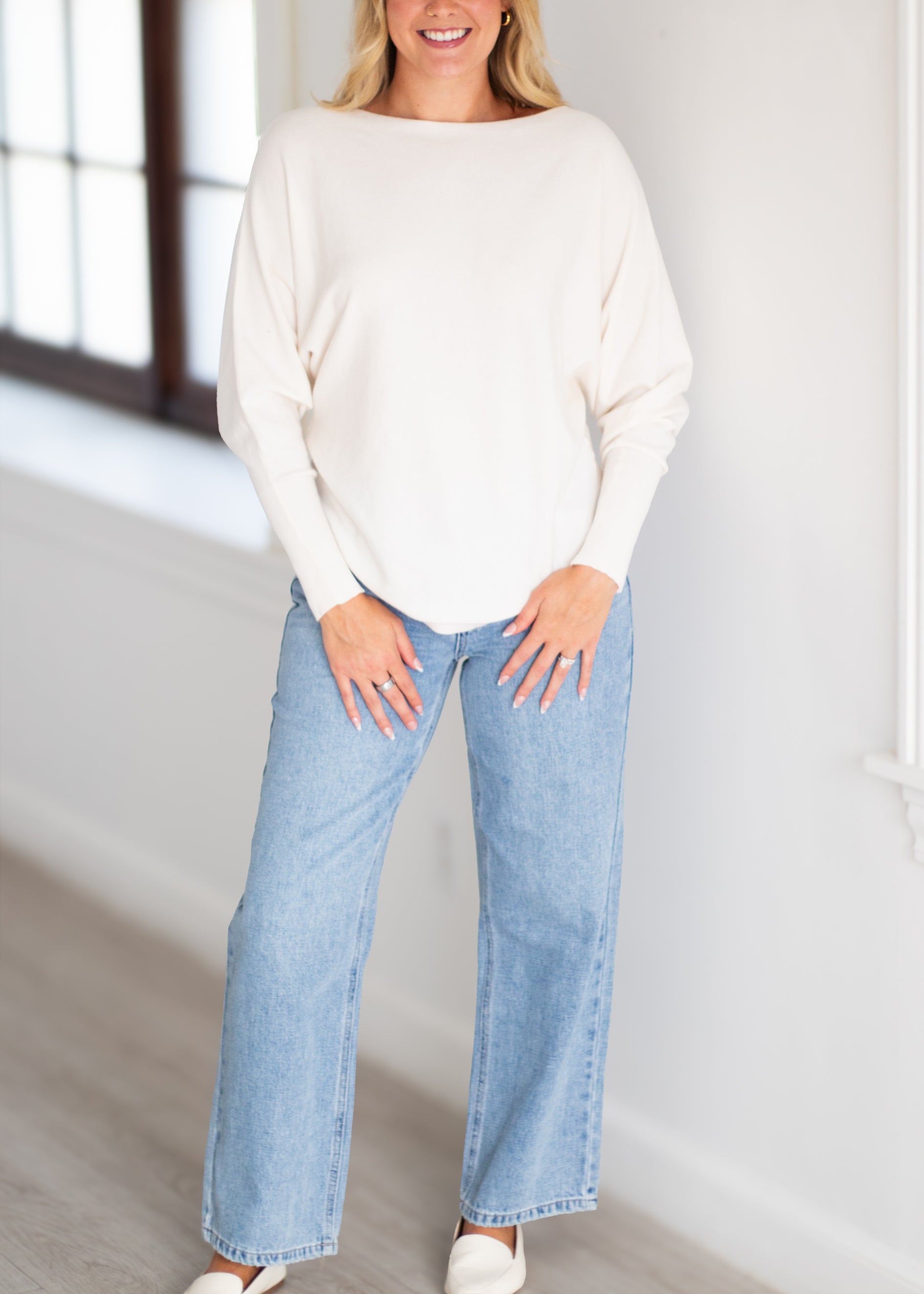 A woman wearing a cream-colored boatneck knit sweater and light-wash wide-leg jeans, standing in a bright room with a window.