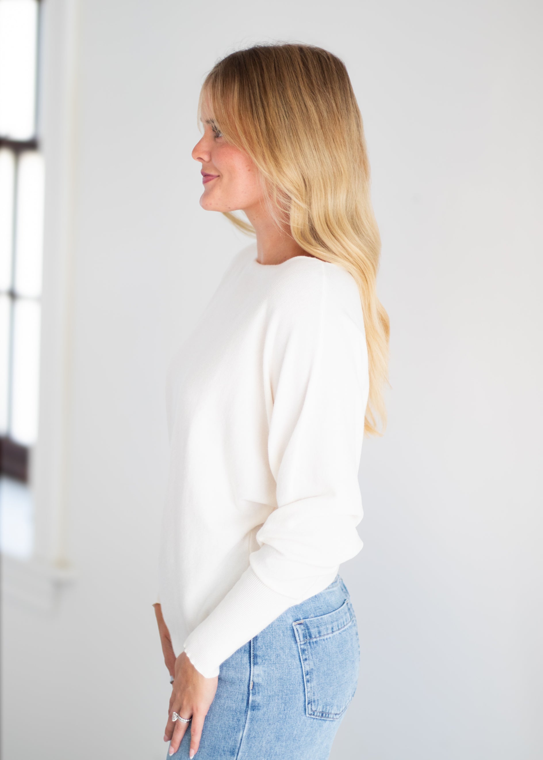 A woman with blonde hair is shown in profile wearing a cream-colored long-sleeve knit sweater and light blue jeans against a bright white background.