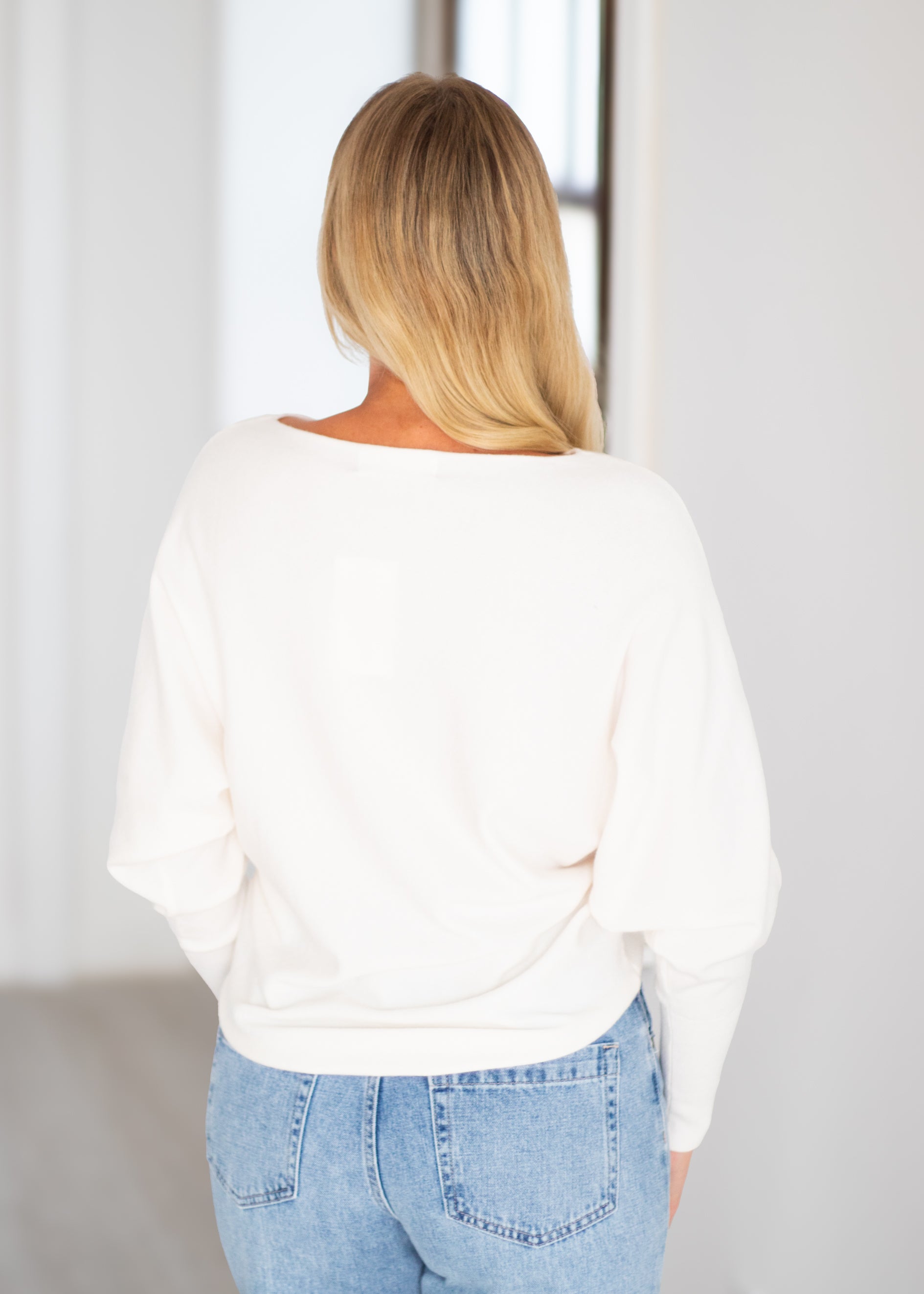 A woman viewed from behind wearing a cream-colored, wide-neck long-sleeve top and light blue jeans in a bright indoor setting.