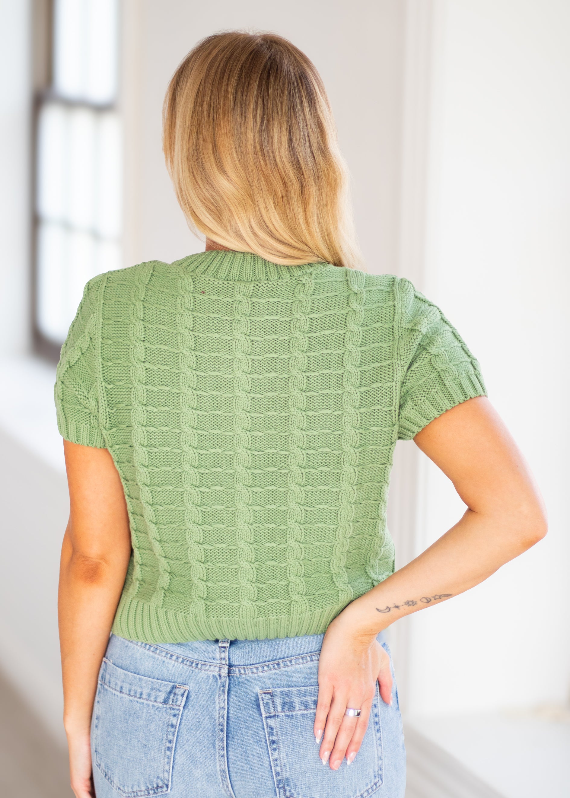 A woman viewed from behind wearing a sage green short-sleeve cable-knit sweater and light blue jeans in a bright indoor setting.