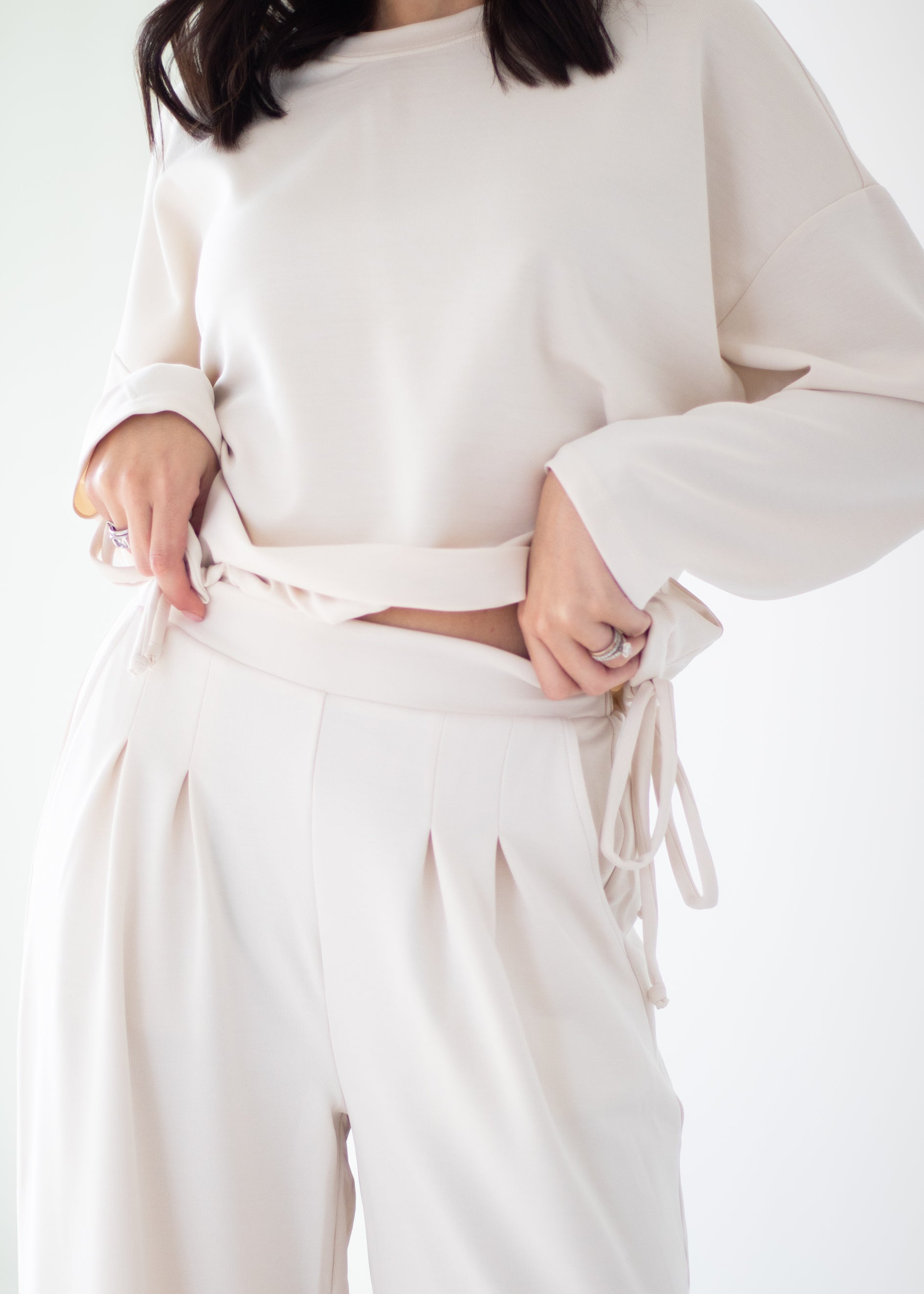 A person wearing a matching cream-colored lounge set consisting of a cropped long-sleeve top and high-waisted pleated trousers with a tie waist.