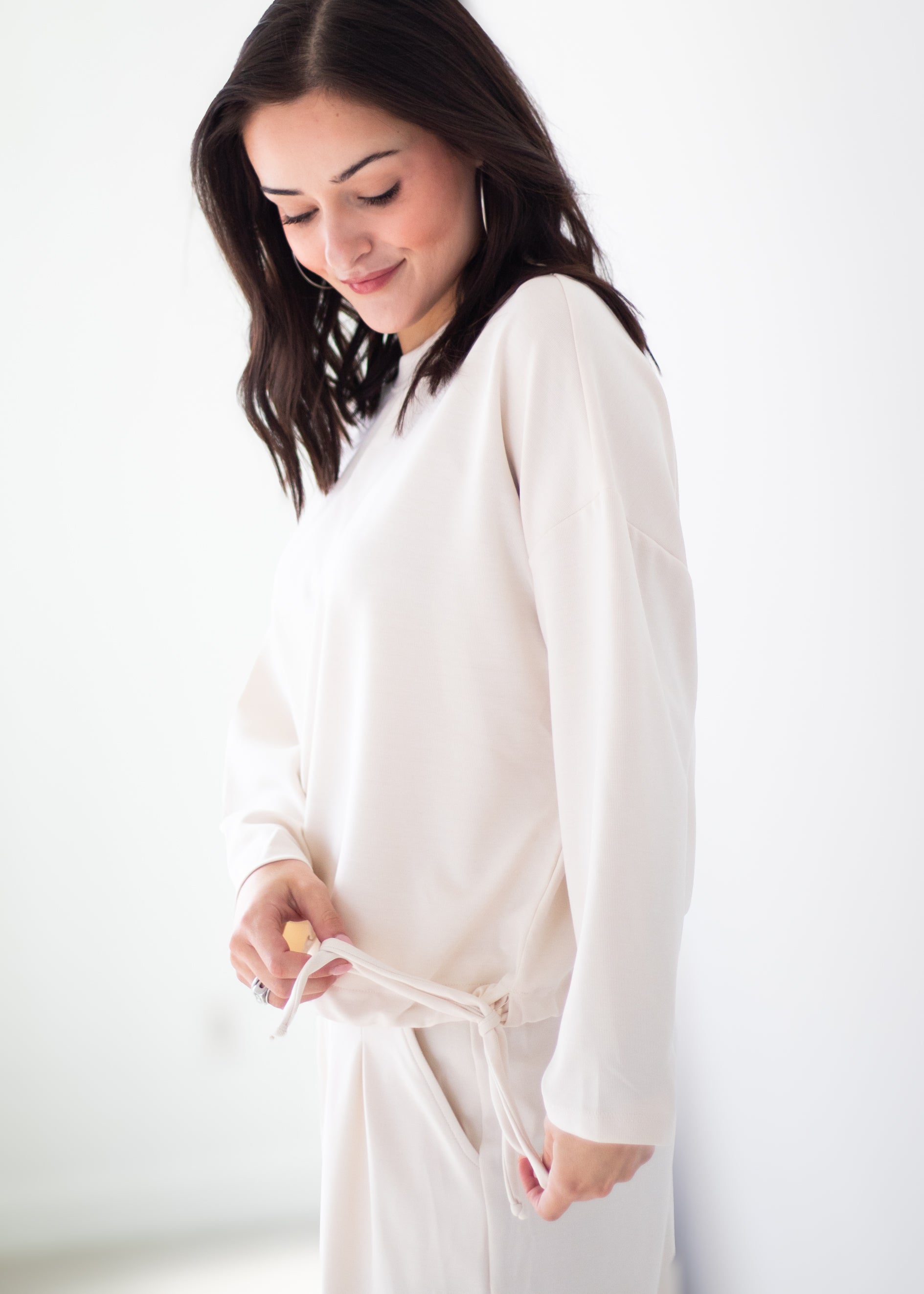 A woman wearing a cream-colored long-sleeve tunic top and matching pants, standing against a plain white wall.
