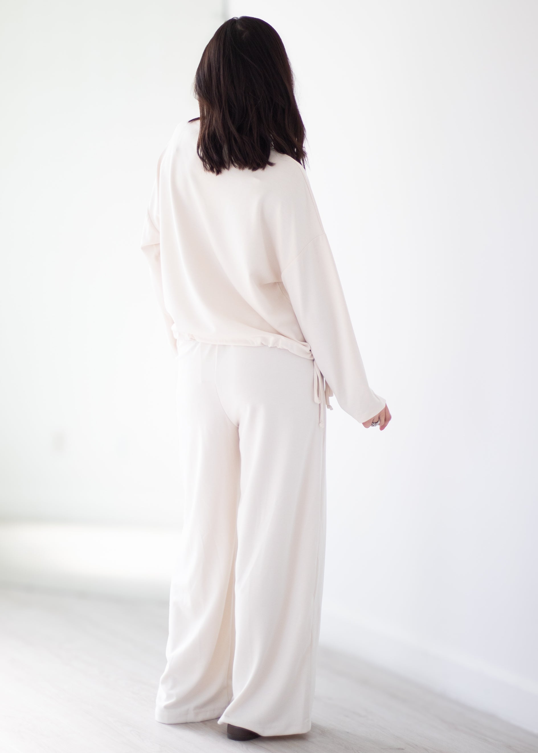 A woman viewed from behind wearing a matching cream-colored lounge set consisting of a long-sleeve top and wide-leg pants, standing in a bright, minimalist room.