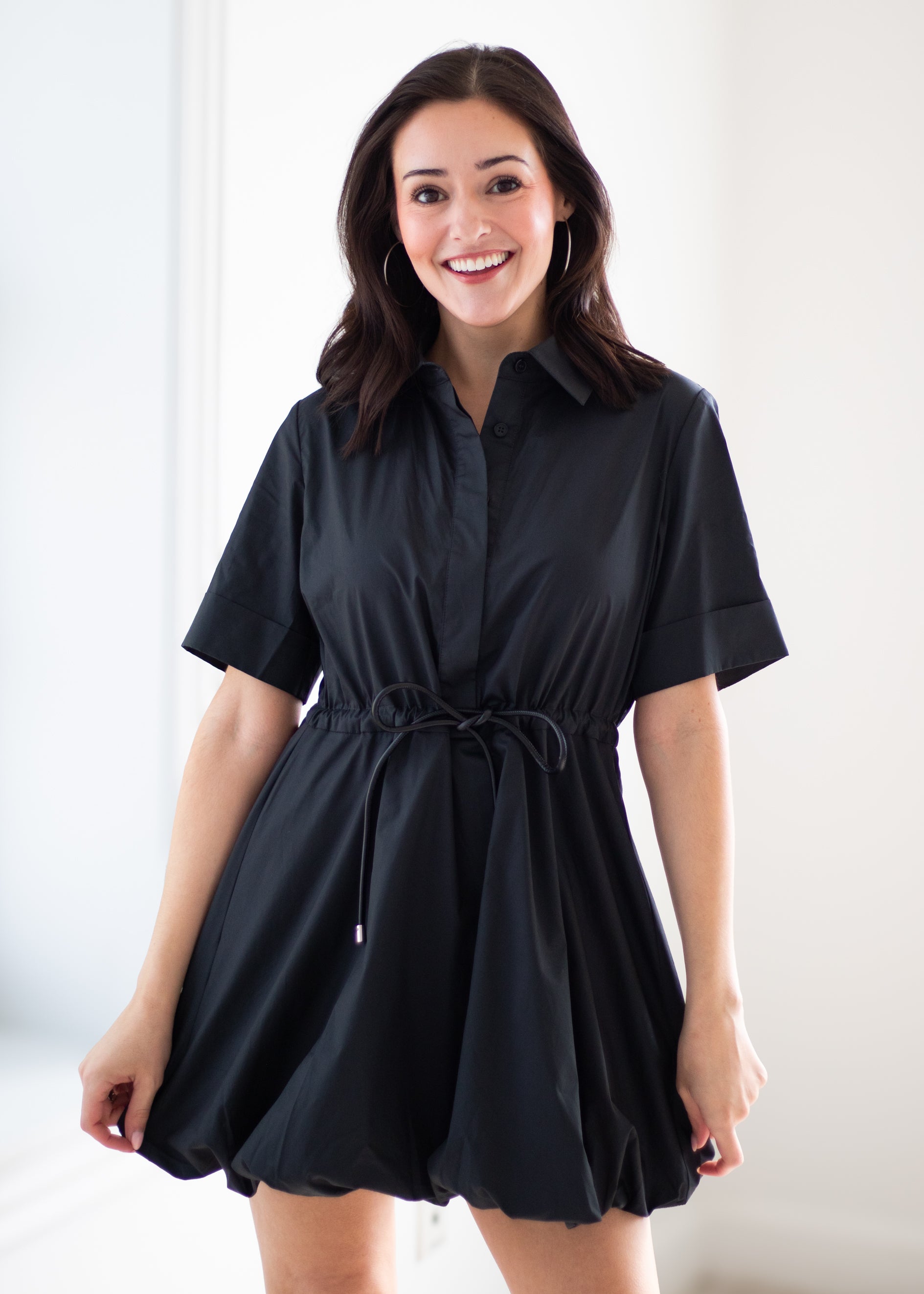 A woman modeling a black short-sleeve collared shirt dress with a drawstring waist, standing against a bright white background.