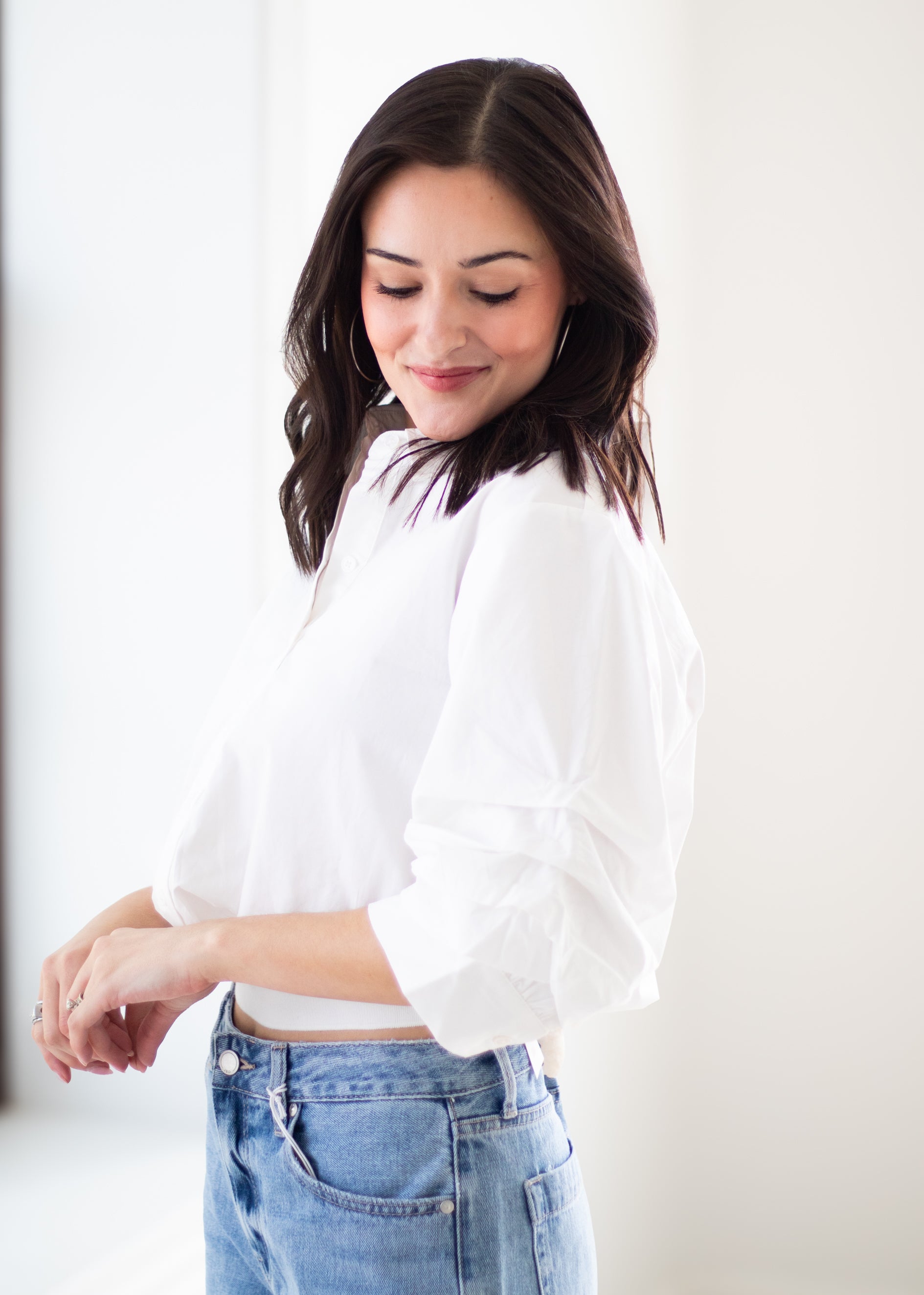 A woman wearing a white long-sleeve blouse with gathered, balloon-style sleeves and light blue jeans, standing against a bright white background.