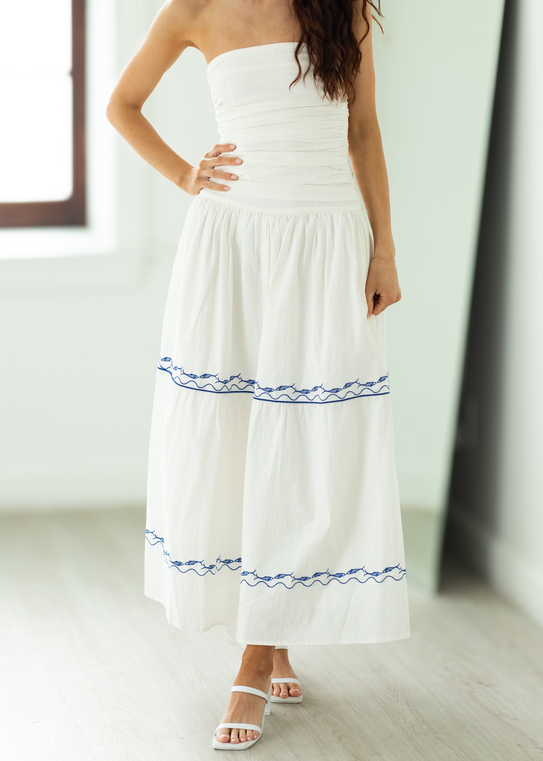 Woman wearing a white strapless dress with blue embroidery in a minimal indoor setting.