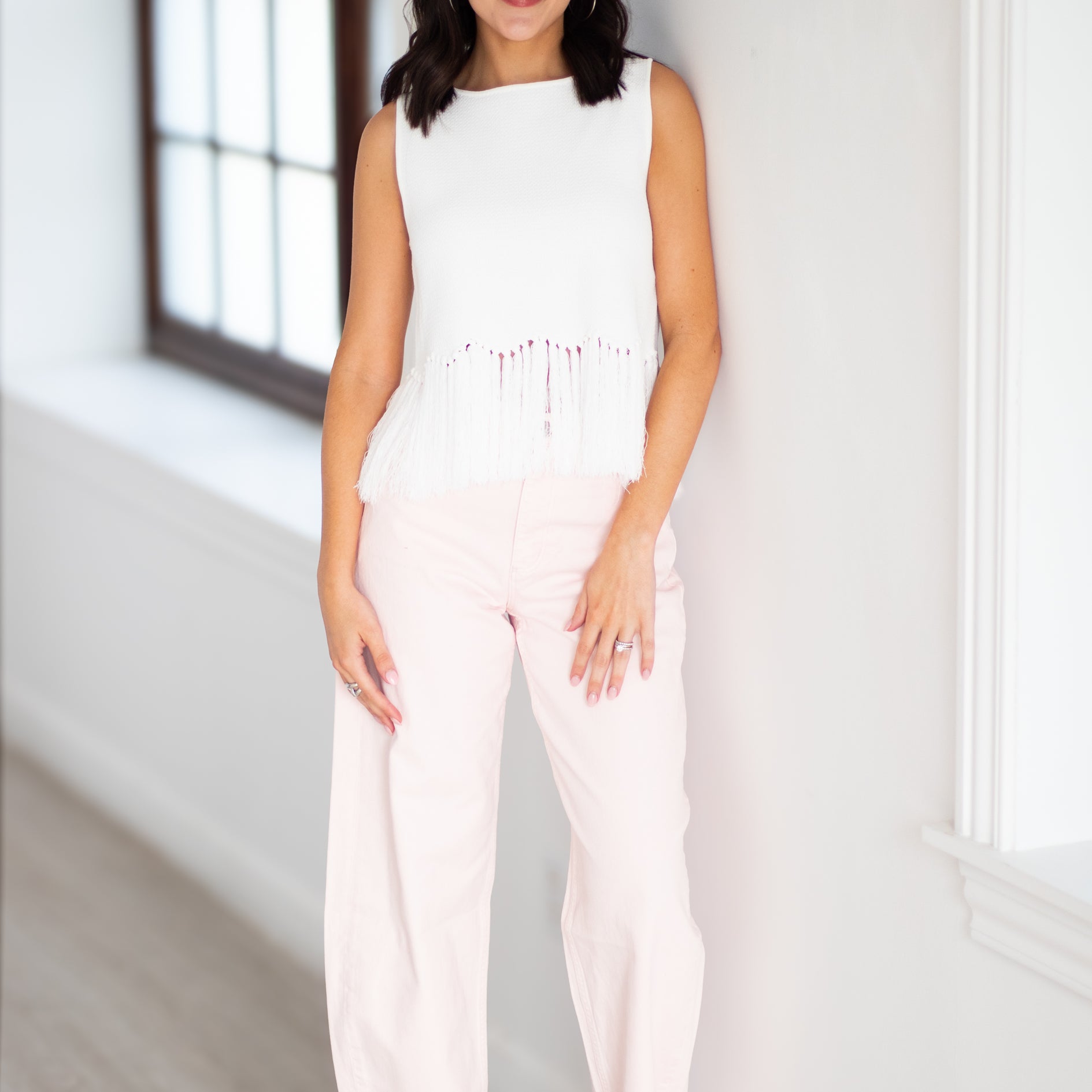 A woman modeling a sleeveless white top with fringe detailing and light pink wide-leg pants, standing in a bright indoor setting with white walls and a window.