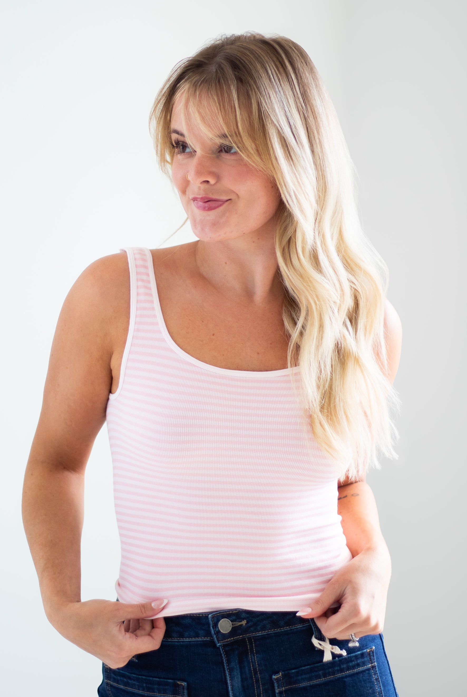 A woman with blonde hair wearing a light pink and white striped ribbed tank top and dark blue jeans, posing against a light gray studio background.