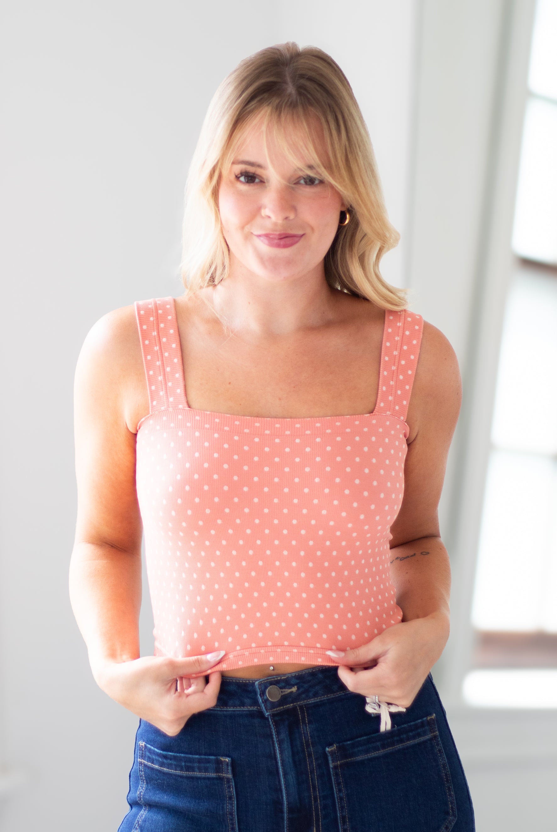 A woman wearing a coral pink and white polka dot tank top with a square neckline, paired with dark blue jeans, standing in a brightly lit room.