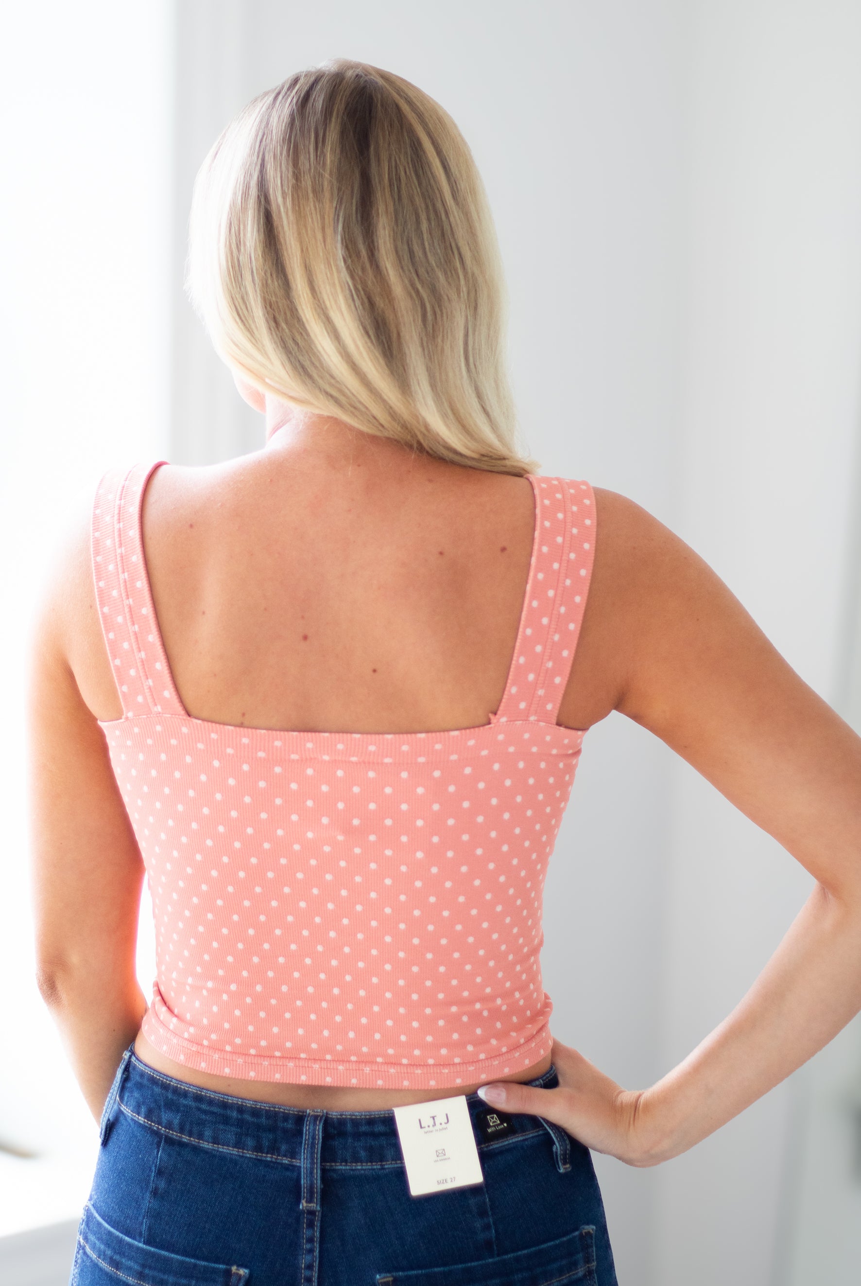 A woman viewed from behind wearing a coral pink tank top with small white polka dots and blue jeans, standing in a bright indoor setting.