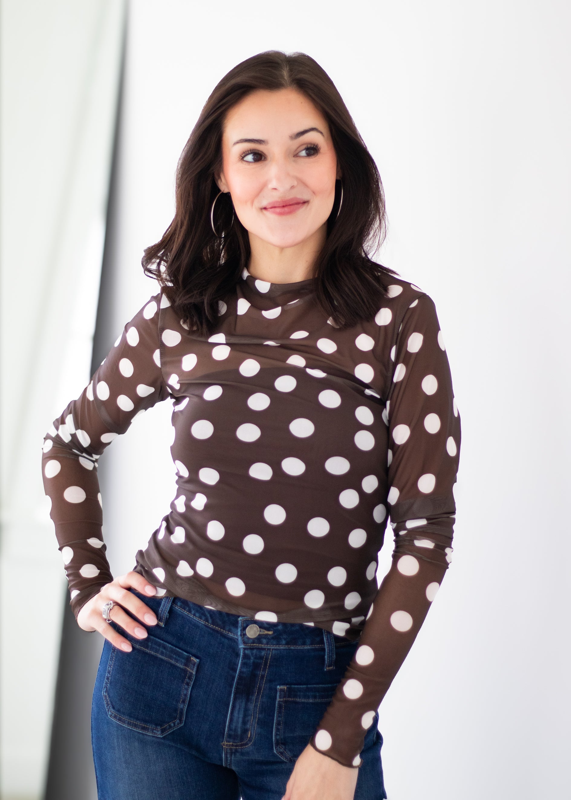 A woman wearing a brown long-sleeve mock neck top with large white polka dots, paired with blue jeans, standing against a white studio background.