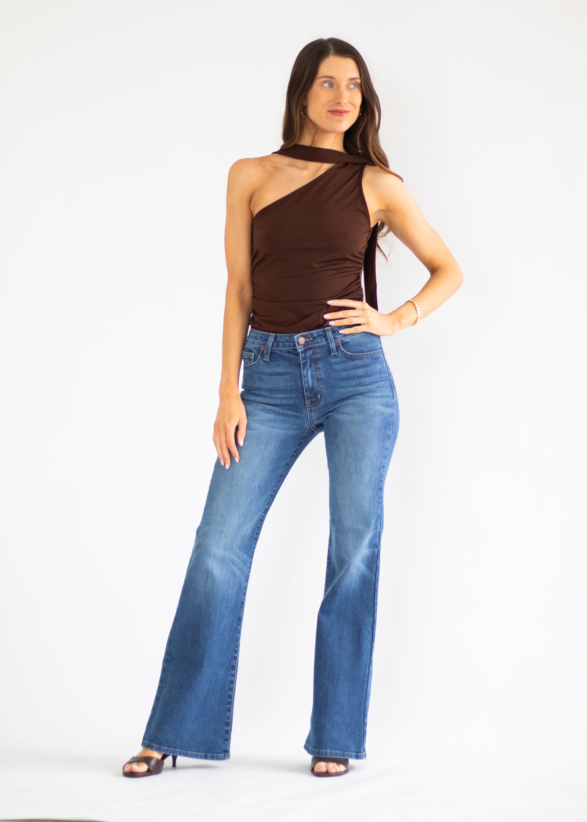 A woman modeling a brown one-shoulder halter top and blue high-waisted flare jeans against a plain white studio background.