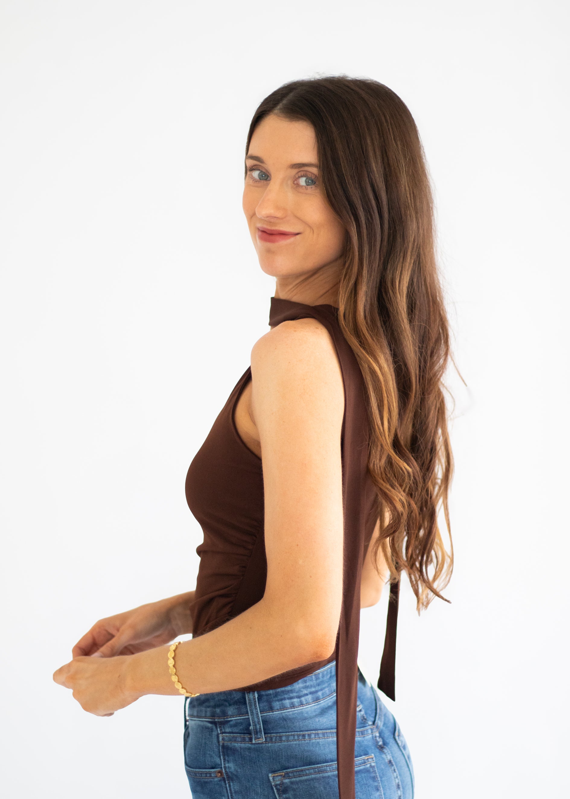 A woman with long brown hair is shown from the side/back wearing a sleeveless dark brown top with a long tie detail and blue jeans, photographed against a plain white studio background.