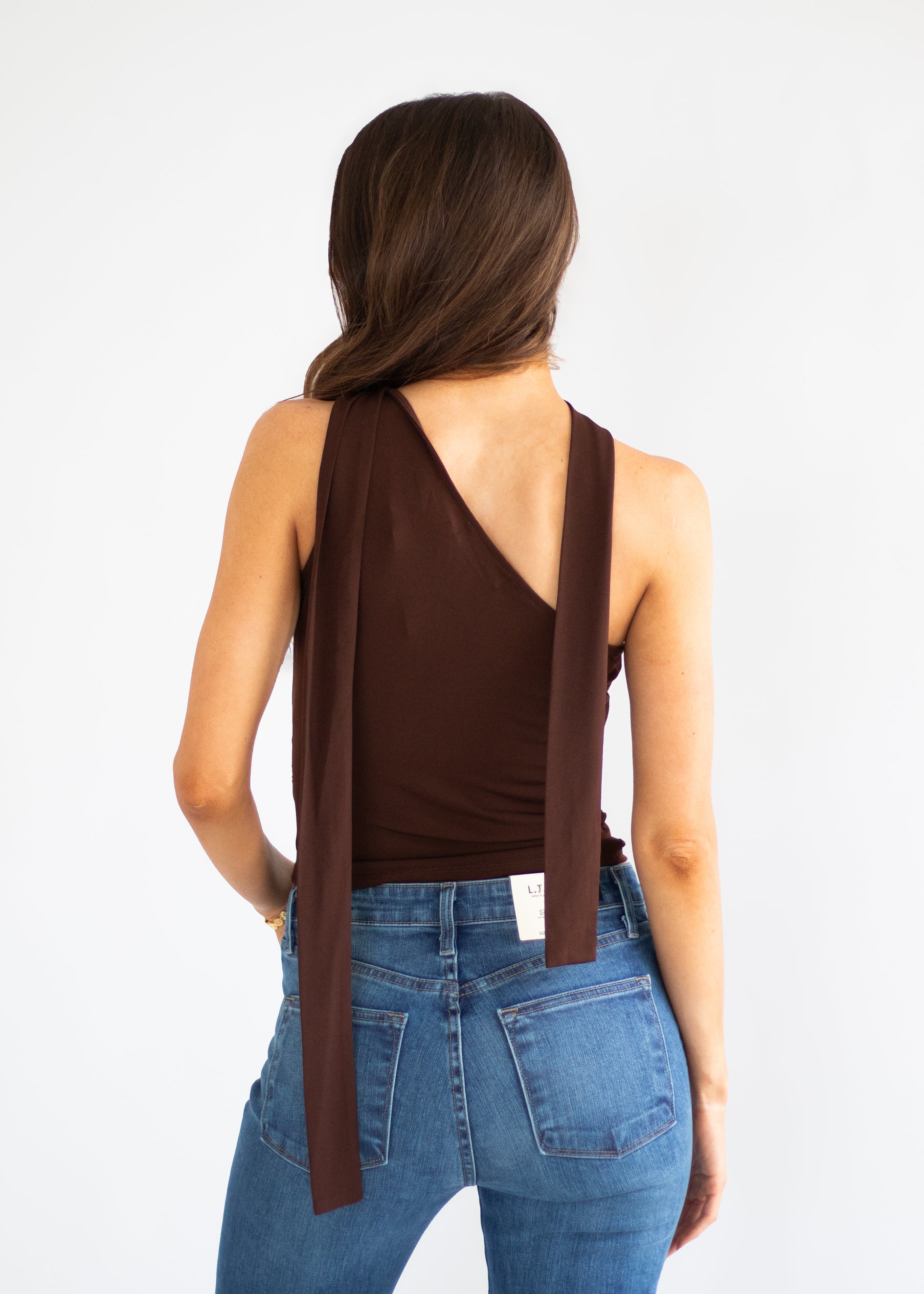 A woman shown from the back wearing a dark brown halter-style top with long tie straps and blue jeans, against a plain white studio background.