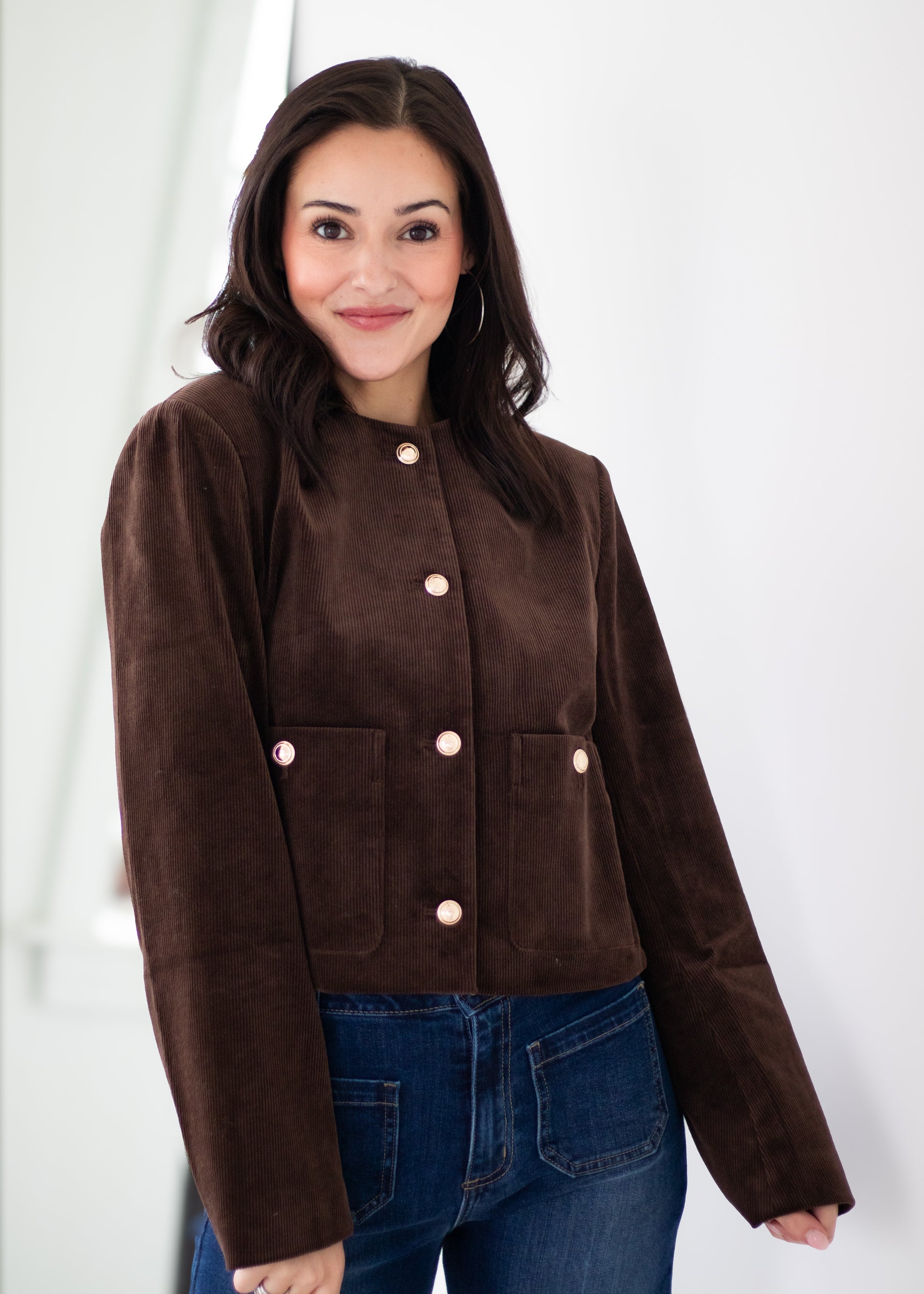 A woman wearing a cropped brown corduroy jacket with light pink buttons, paired with blue jeans, standing against a white background.