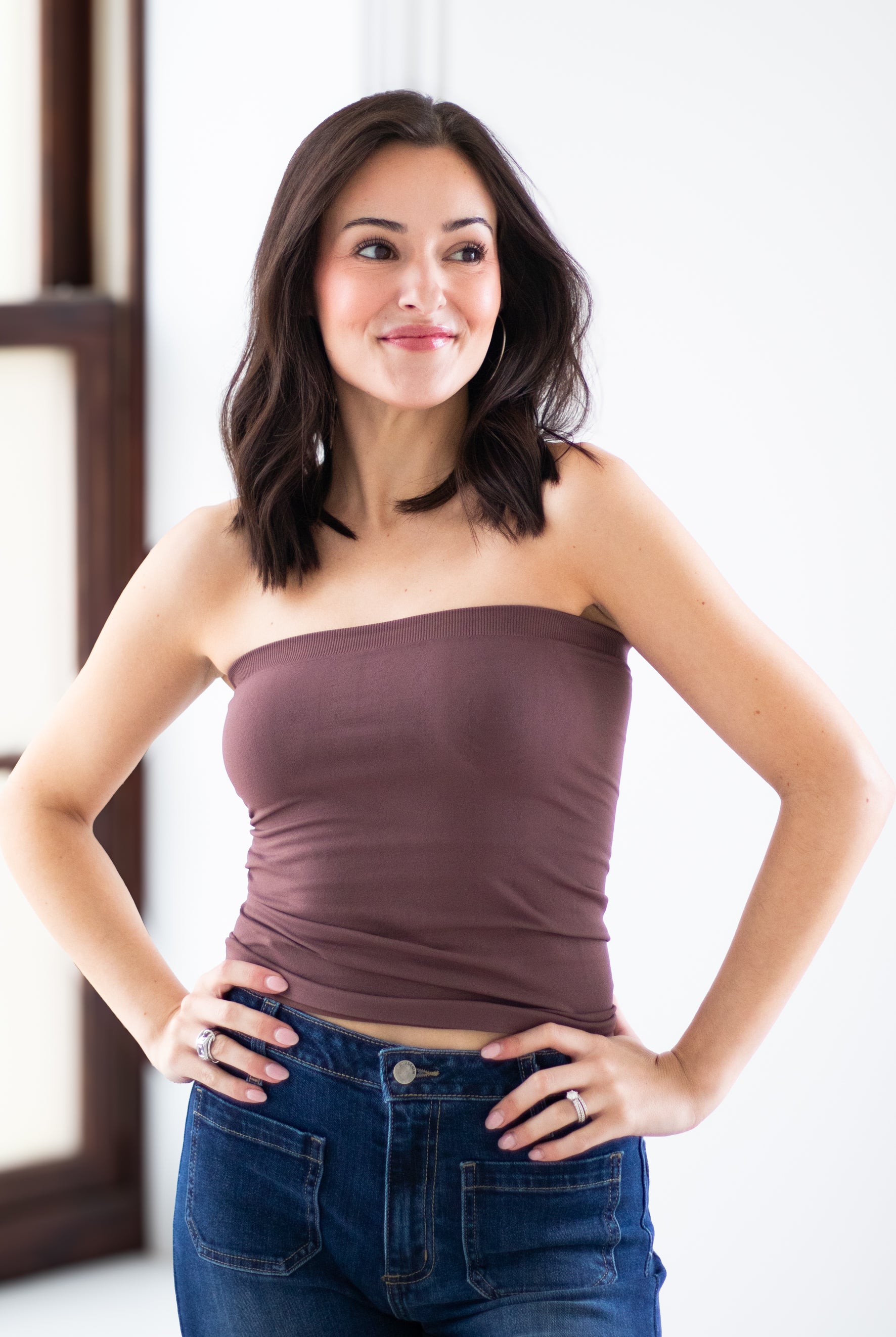 A woman with dark shoulder-length hair wearing a strapless mauve-brown tube top and blue jeans, posing with her hands on her hips against a bright white indoor background.