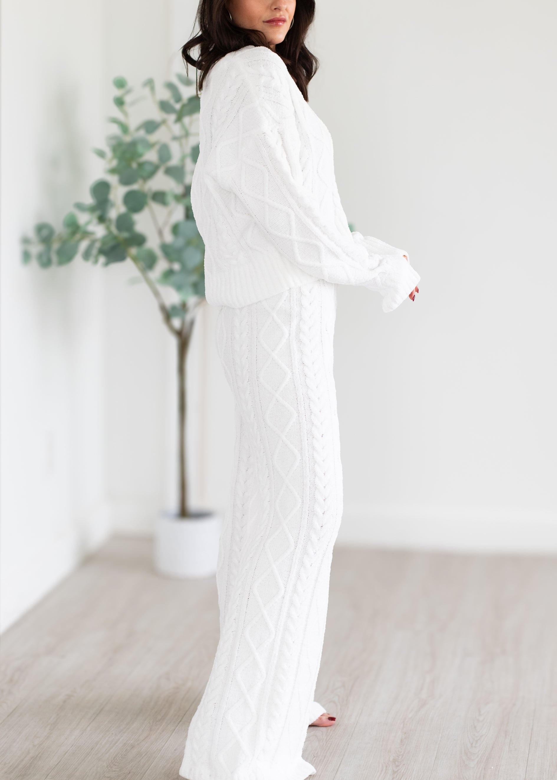 Woman wearing a white dress standing in a minimalistic room with a plant in the background.