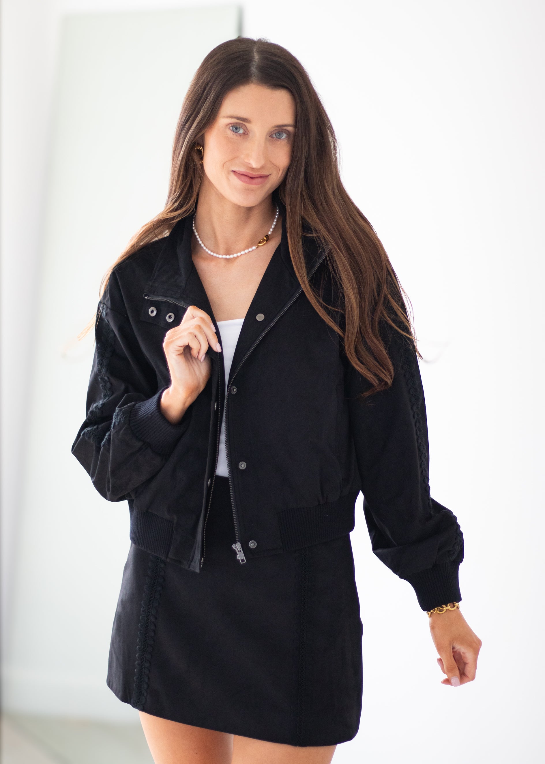 A woman modeling a black denim jacket and matching black denim mini skirt over a white top, standing against a bright white background.