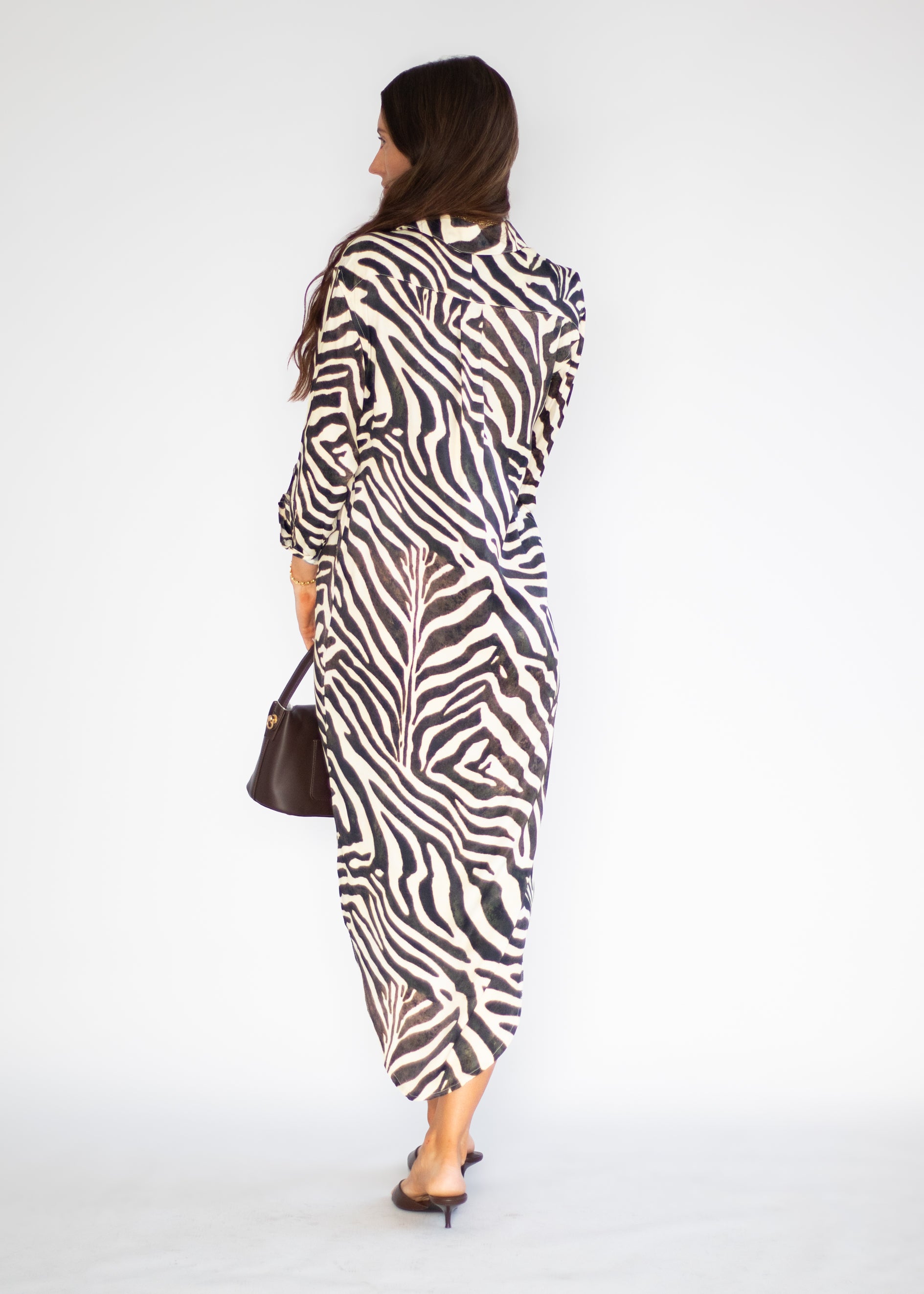 A woman is shown from behind wearing a long-sleeved zebra-print midi dress with a shawl collar. She is holding a small dark brown handbag and wearing black pointed-toe flats, set against a plain white studio background.