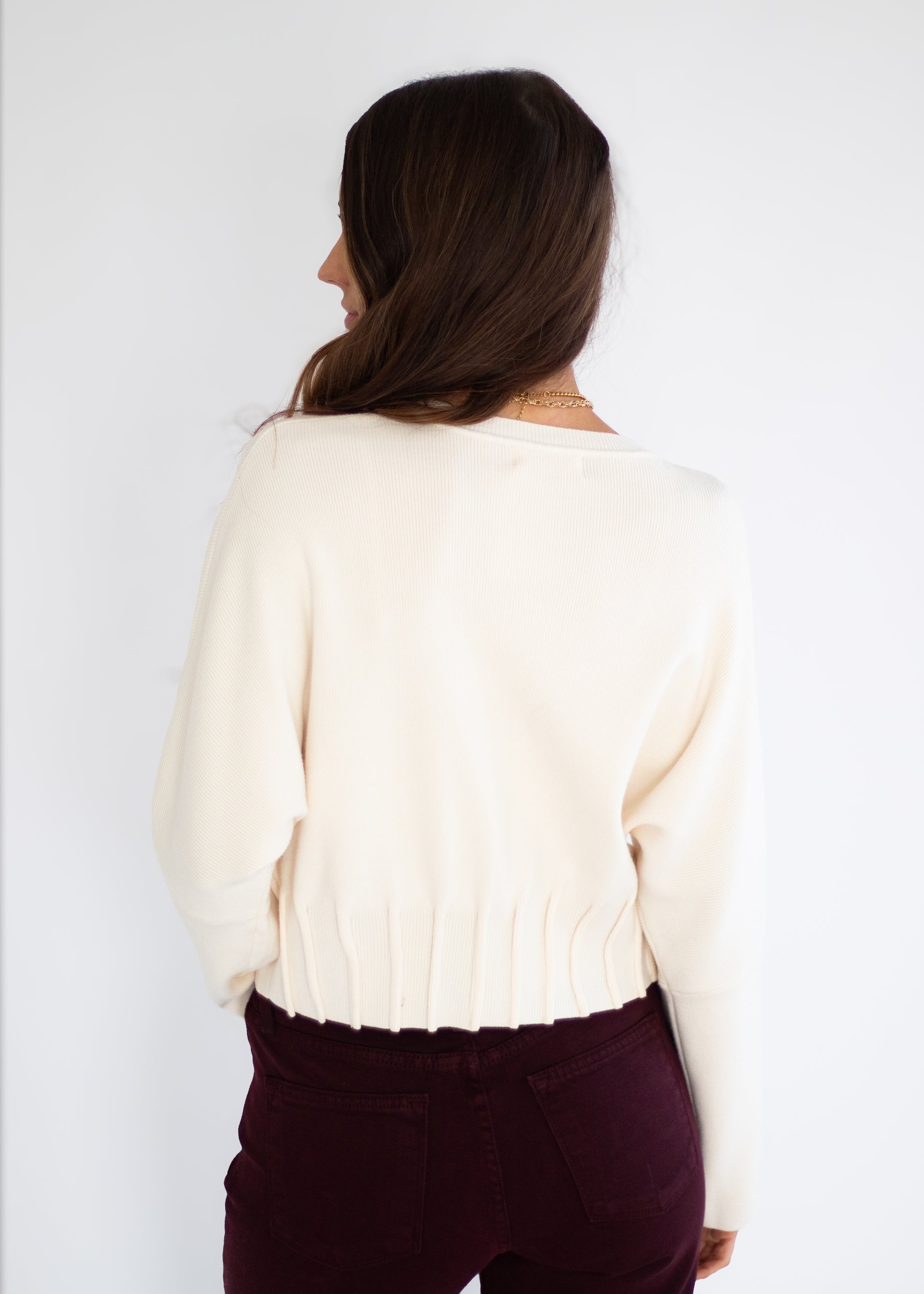 A woman shown from behind wearing a cream-colored knit sweater and dark burgundy pants against a plain white background.