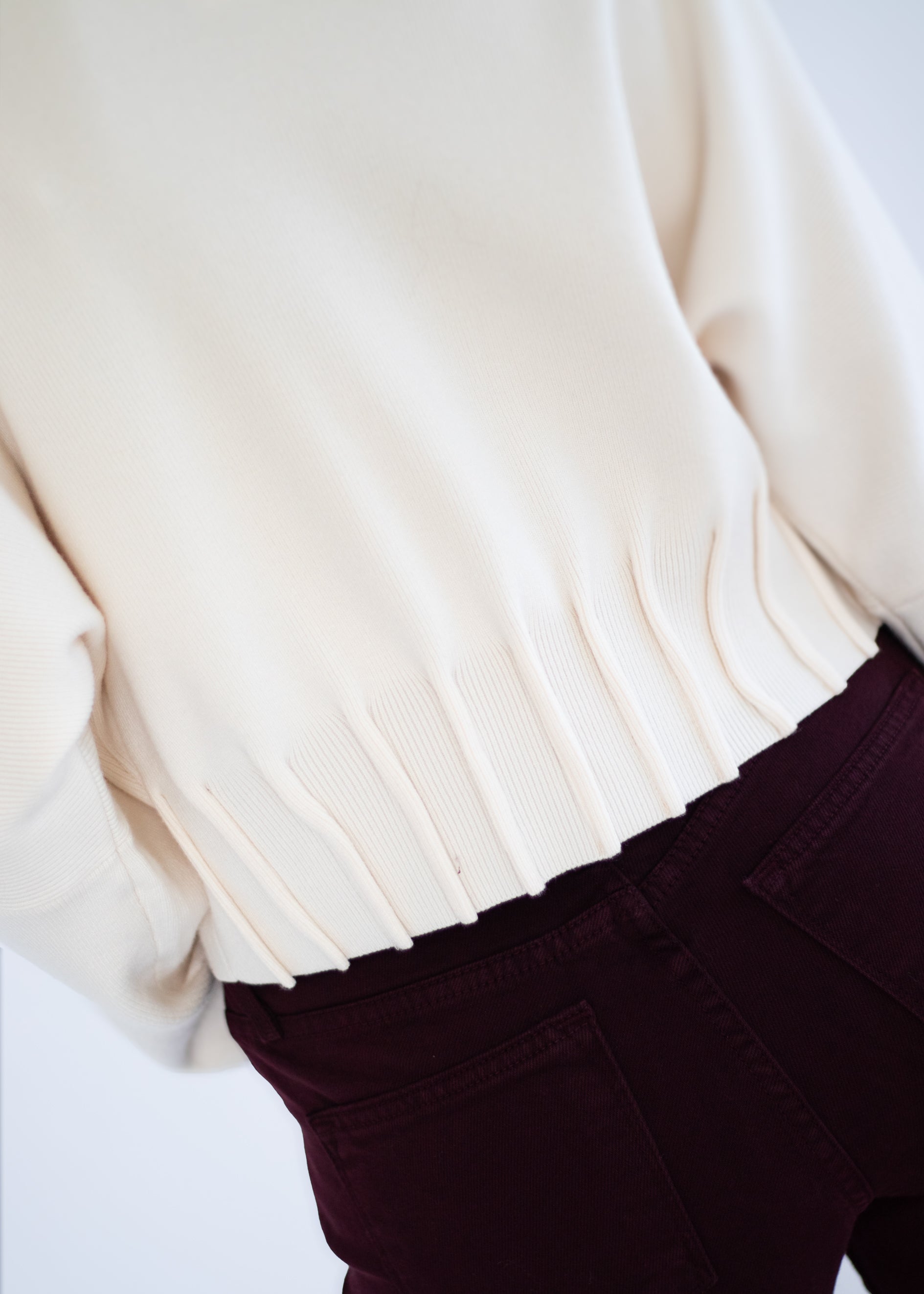 Close-up of a person wearing a cream knit sweater with a ribbed hem and dark burgundy pants against a plain light background.