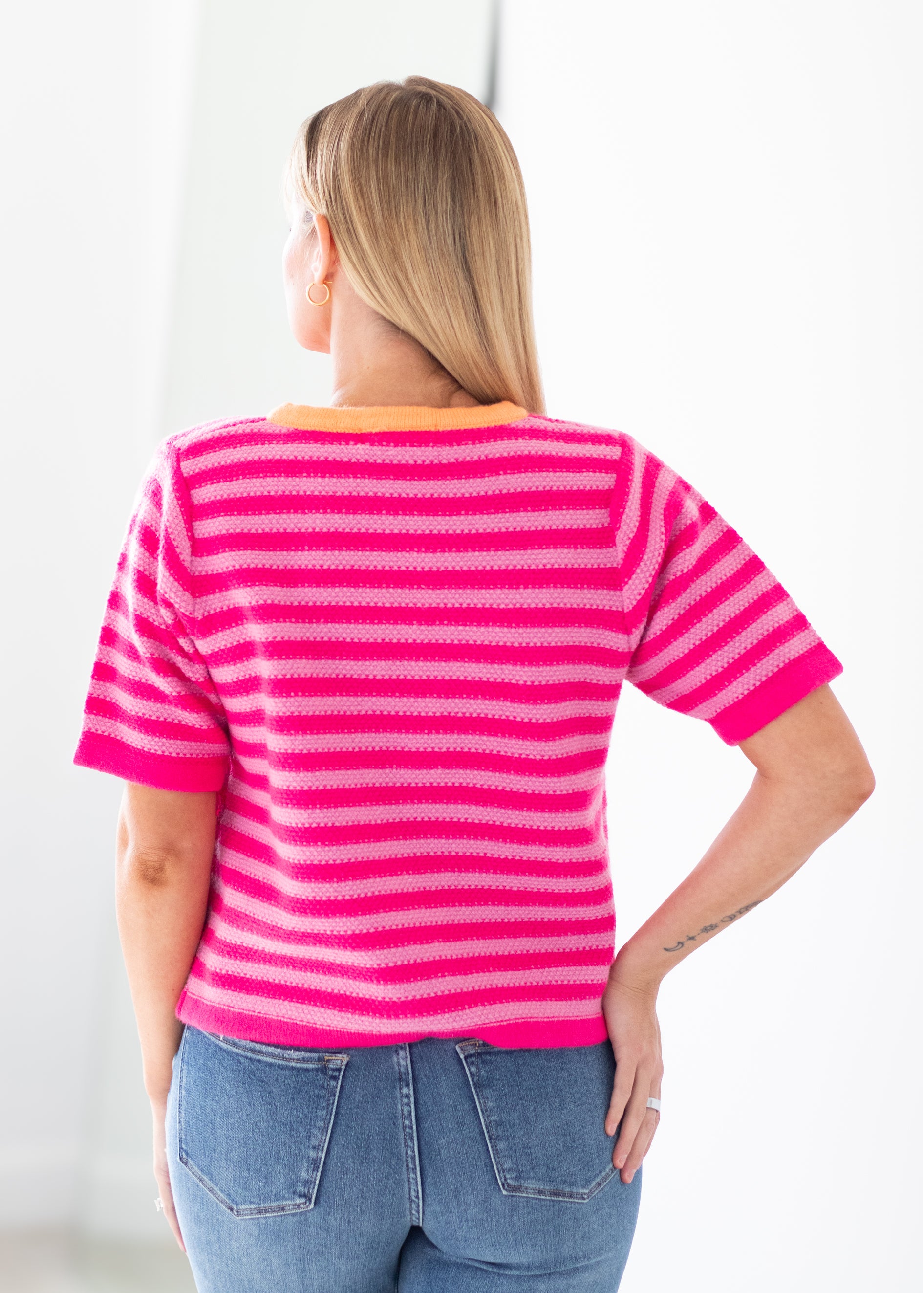 A woman viewed from behind wearing a bright pink and magenta striped short-sleeve knit top with an orange collar, paired with blue jeans, against a clean white background.