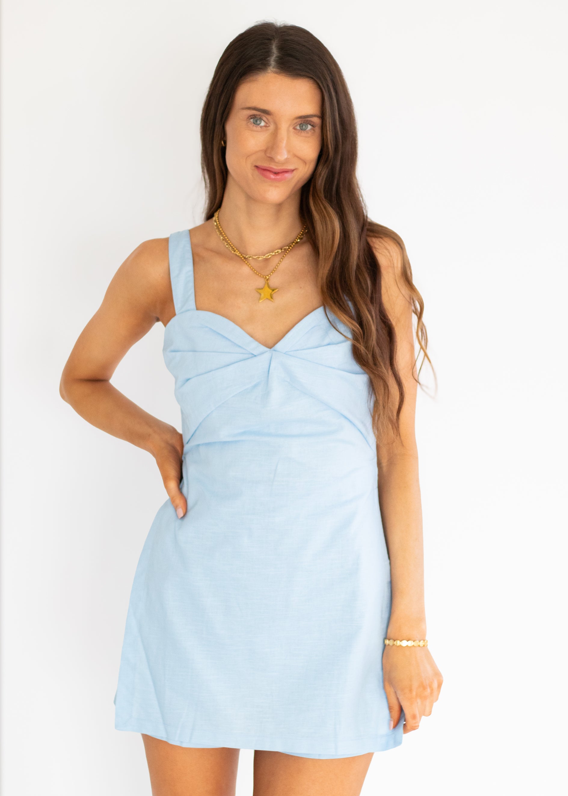 A woman modeling a light blue sleeveless mini dress with a sweetheart neckline and gathered front detail, photographed against a plain white studio background.