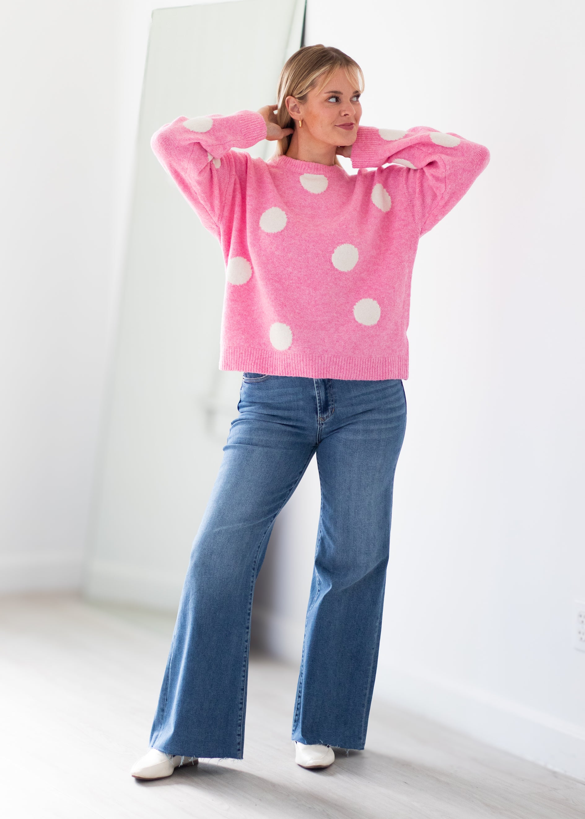A woman modeling a pink knit sweater with large white polka dots, paired with blue flared jeans and white shoes, standing in a bright white room.