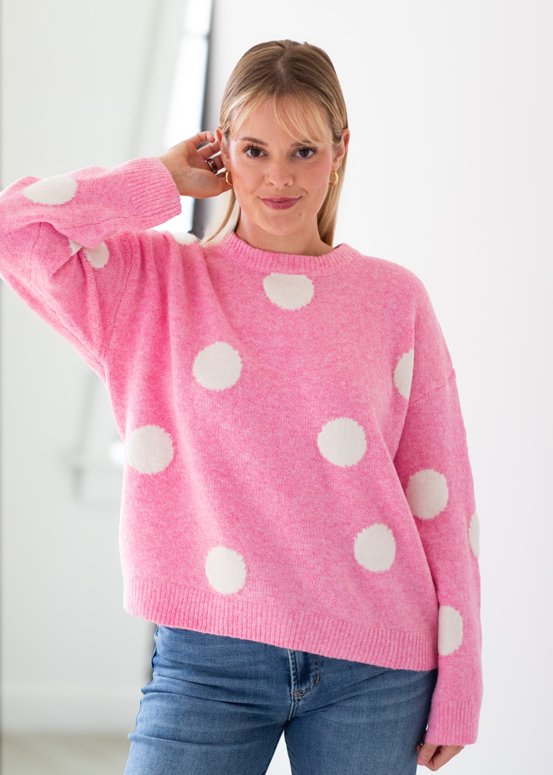 A woman wearing a pink knit sweater with large white polka dots, paired with blue jeans, standing in a bright indoor setting.