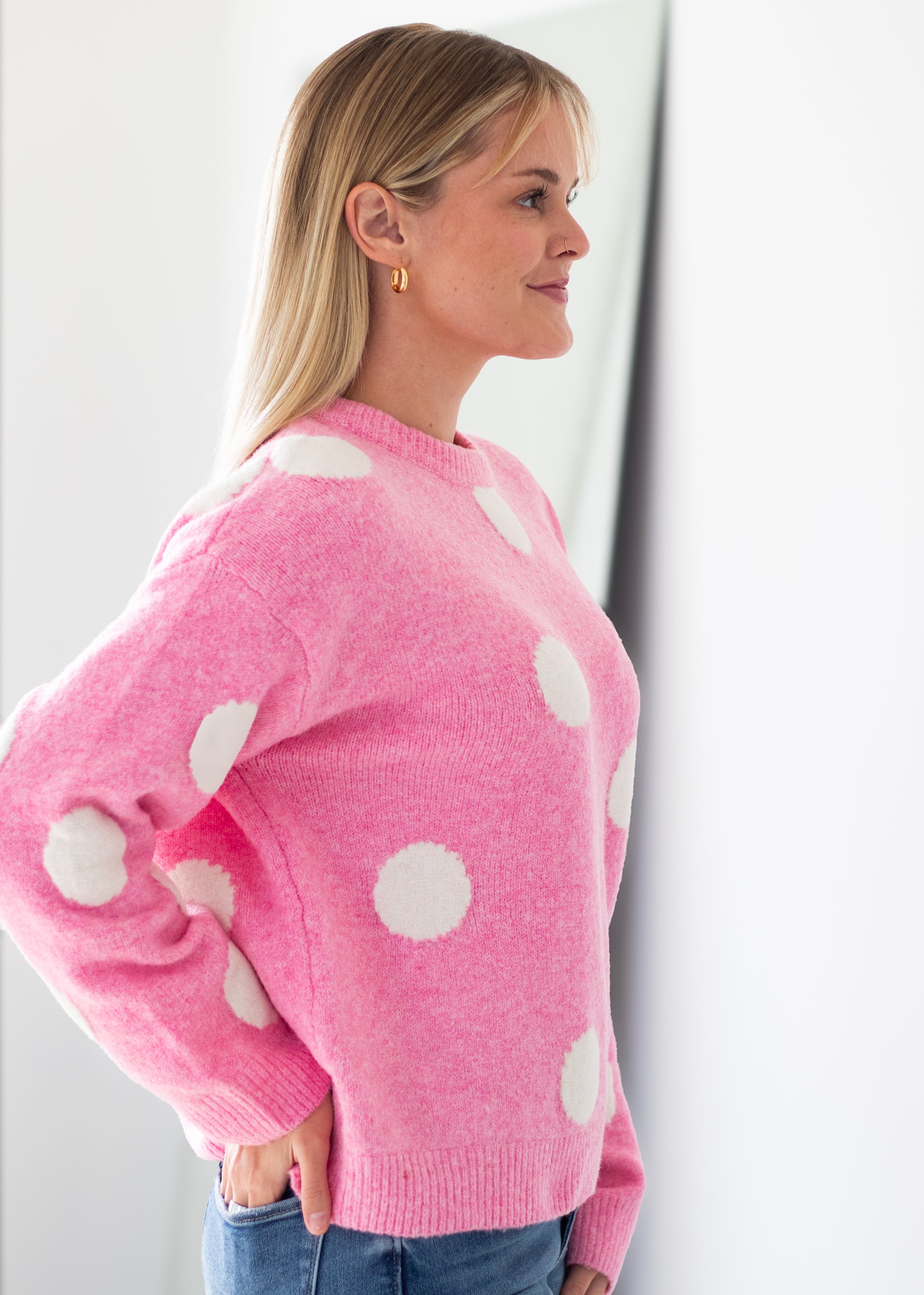 A woman in profile wearing a pink knit sweater with large white polka dots, paired with blue jeans, standing against a bright white wall.