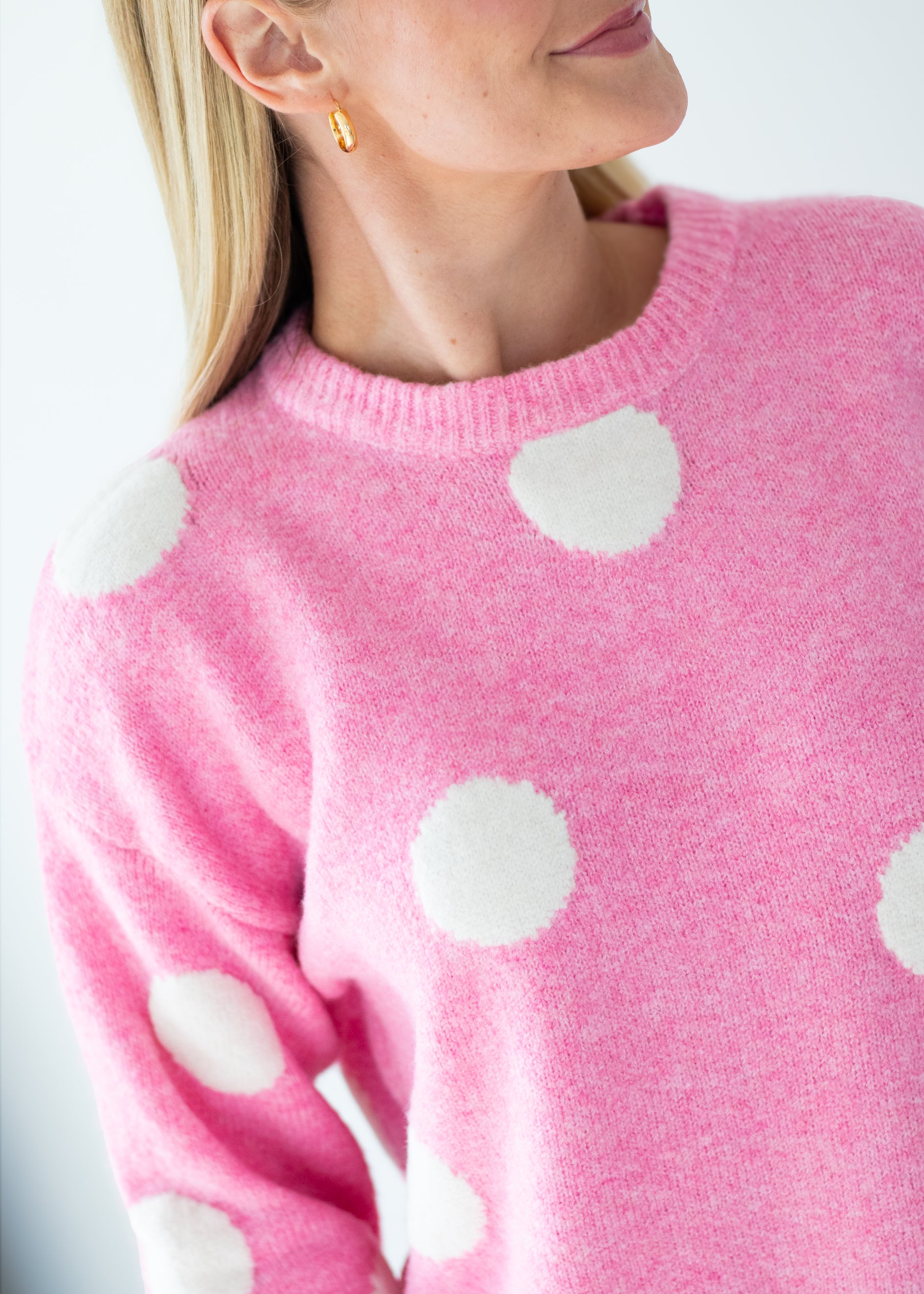 A close-up of a woman wearing a pink knit sweater with large white polka dots, shown from the shoulders to the chest against a light neutral background.