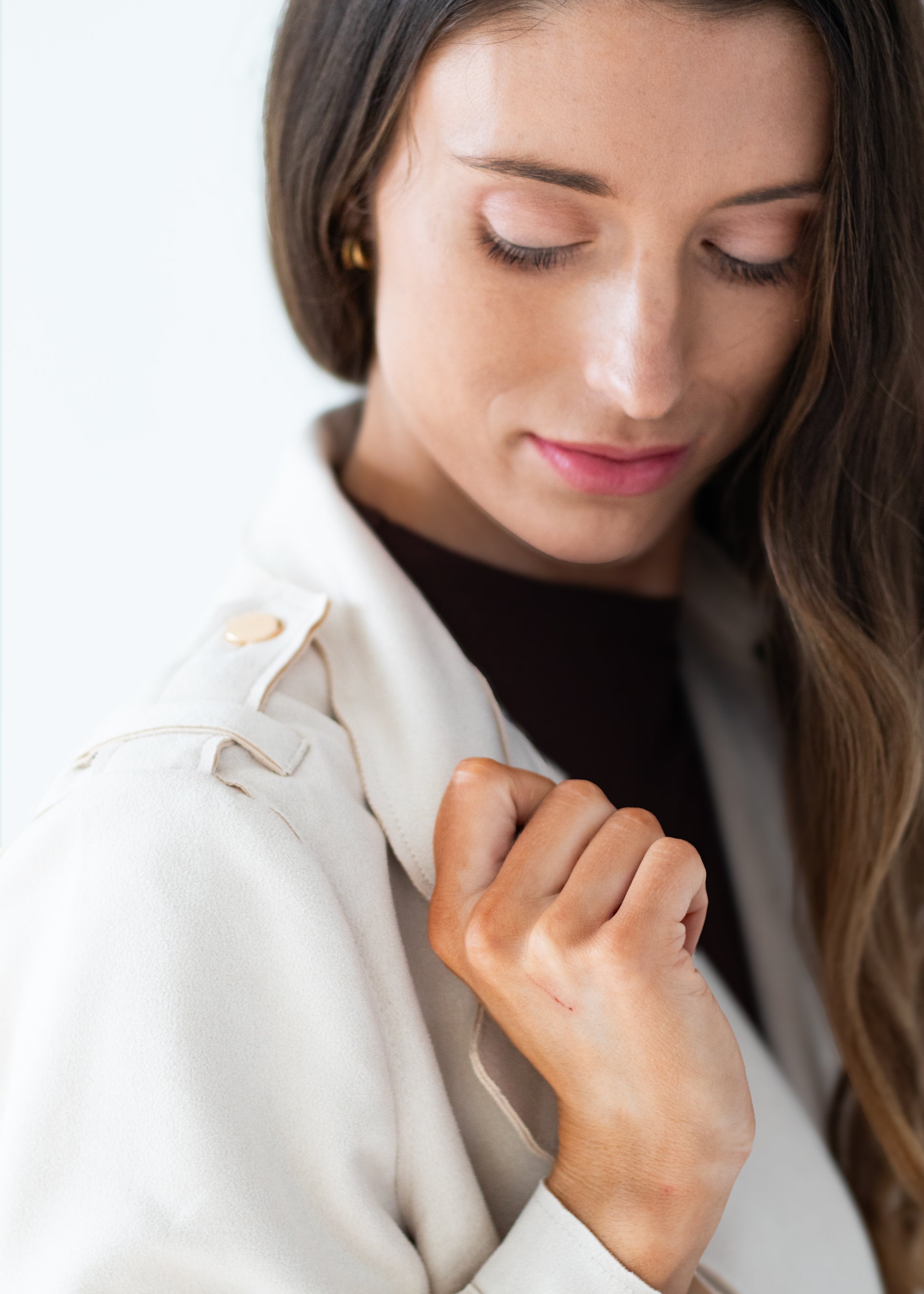 A close-up lifestyle shot of a woman wearing a light cream-colored trench coat over a dark brown top, looking down with a soft expression.