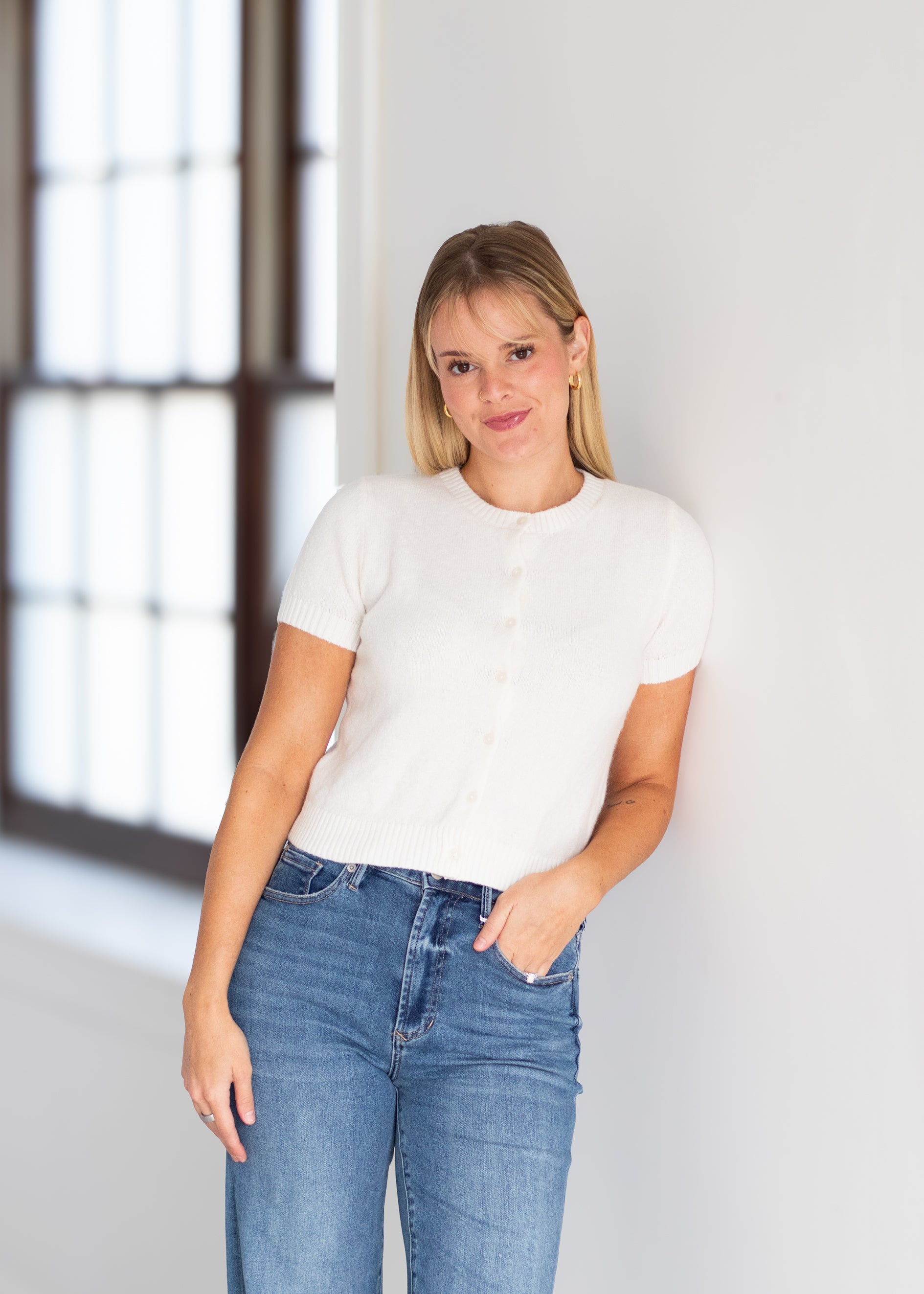 A woman wearing a white short-sleeve button-front knit top and blue jeans, standing indoors against a white wall near a window.