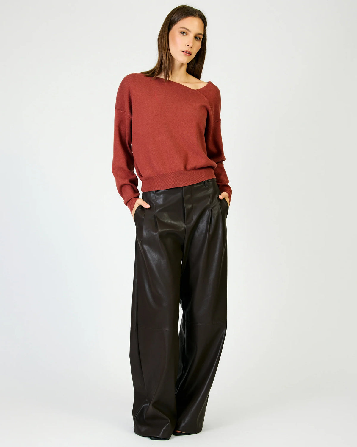 A woman modeling a rust-red long-sleeve knit sweater and black wide-leg faux-leather pants against a light gray studio background.