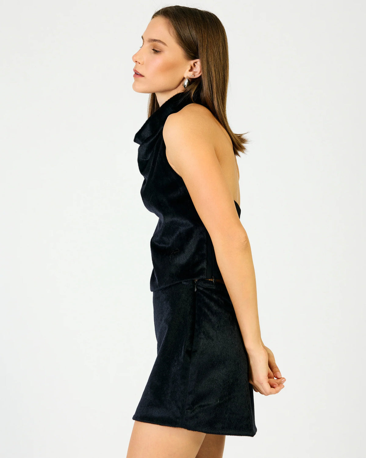 A woman in side profile wearing a sleeveless black top and skirt against a plain light gray studio background.