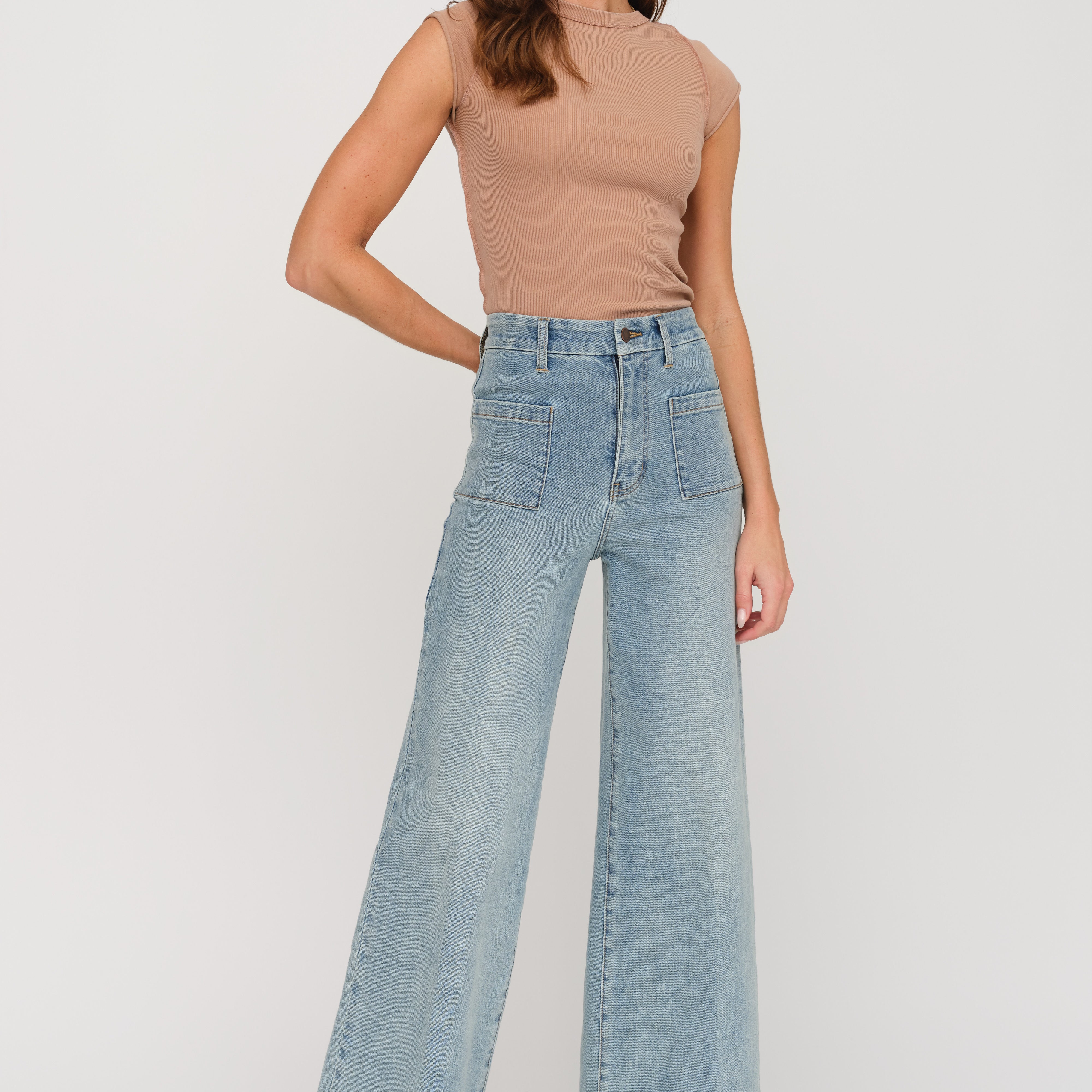 A woman modeling a pair of high-waisted, wide-leg light blue denim jeans with a tan sleeveless top, standing against a plain light gray studio background.