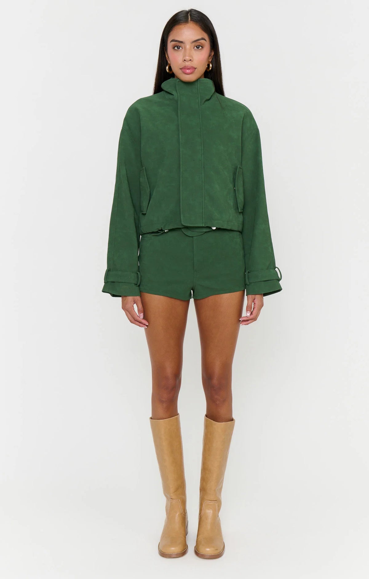 A model stands against a light gray studio background wearing a matching dark green cropped jacket and shorts set, tan knee-high boots, and gold hoop earrings.