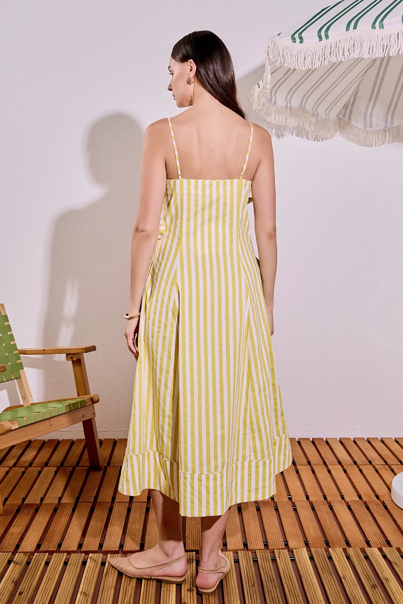 Woman wearing a yellow striped dress standing on a wooden floor with a white wall and chair in the background.