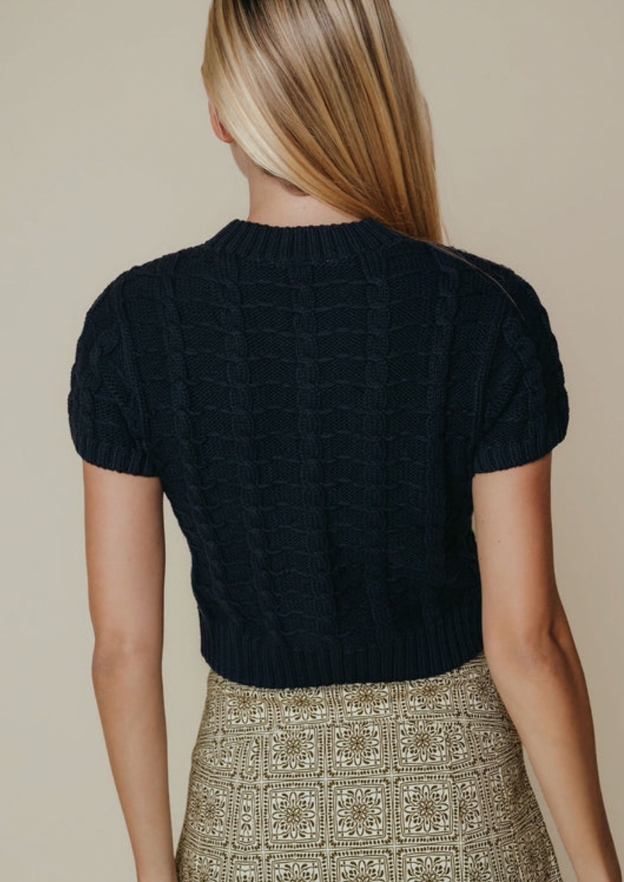 A woman shown from the back wearing a dark navy cable-knit short-sleeve cropped sweater and a beige patterned skirt, against a solid beige background.