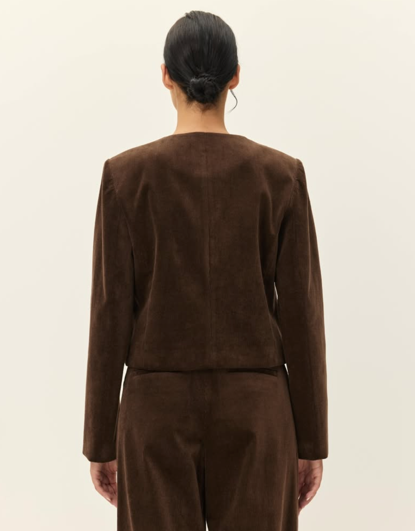 A model shown from the back wearing a dark brown cropped jacket and matching trousers, photographed against a plain off-white studio background.