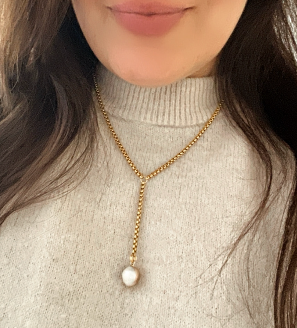 Person wearing a gold necklace with a pearl pendant on a light gray turtleneck sweater.